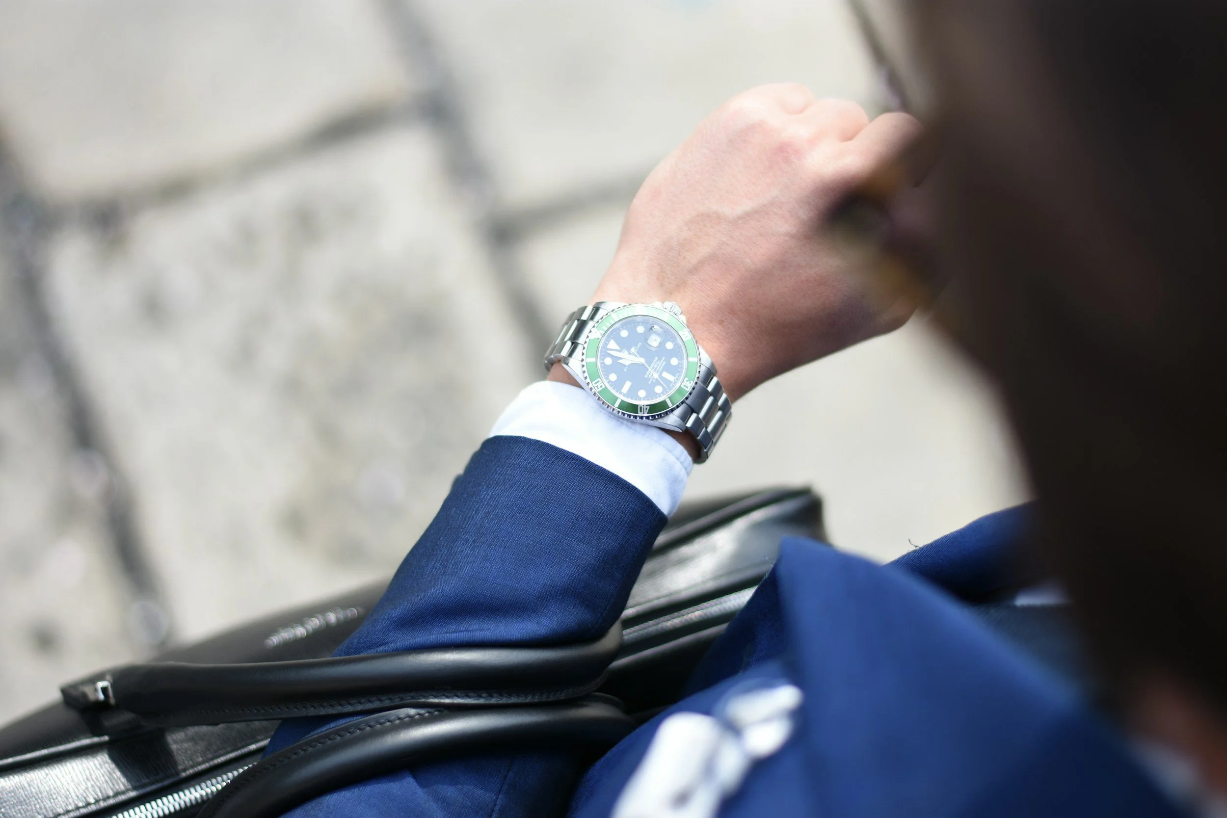 A person in a blue suit wearing a silver wristwatch with a green bezel, sitting in an airport or public transportation setting, with a black bag nearby.