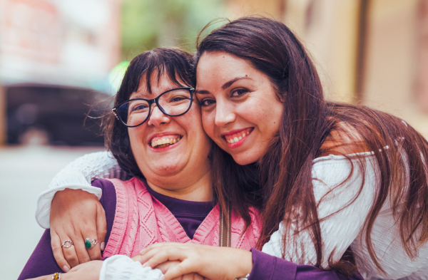 A woman in a wheelchair wearing a purple shirt and pink sweater vest is being embraced by a woman in her twenties with long dark hair and bright red lipstick. She wears an off-white shirt.