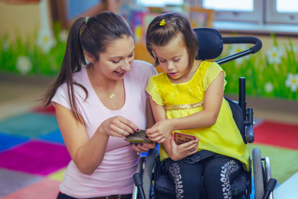 A woman in a pink shirt sits next to a young girl in a wheelchair, wearing yellow. They are looking at flash cards together.