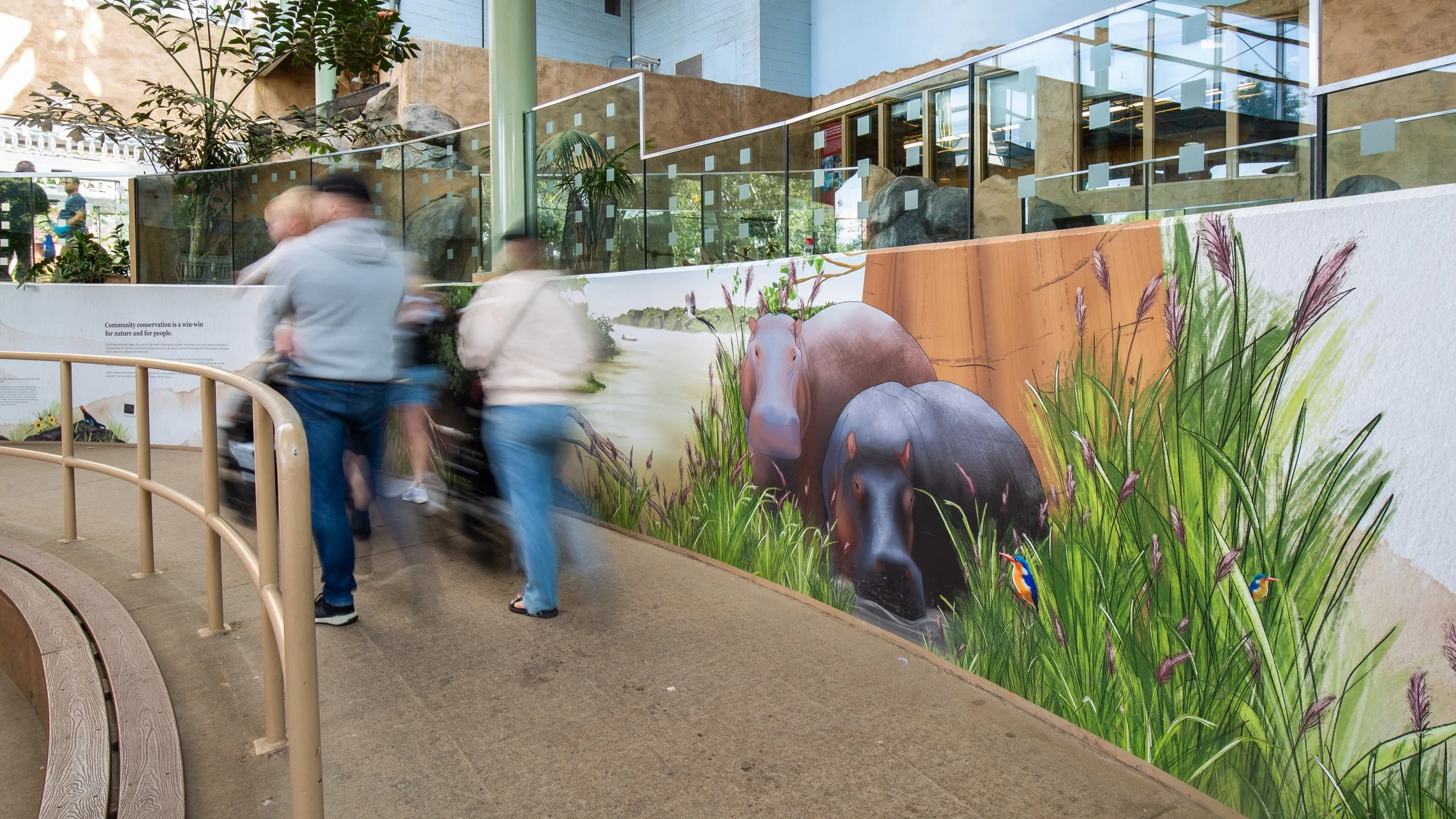 People walking past a colorful mural of hippopotamuses in tall grass with birds, in an indoor nature center or similar setting.