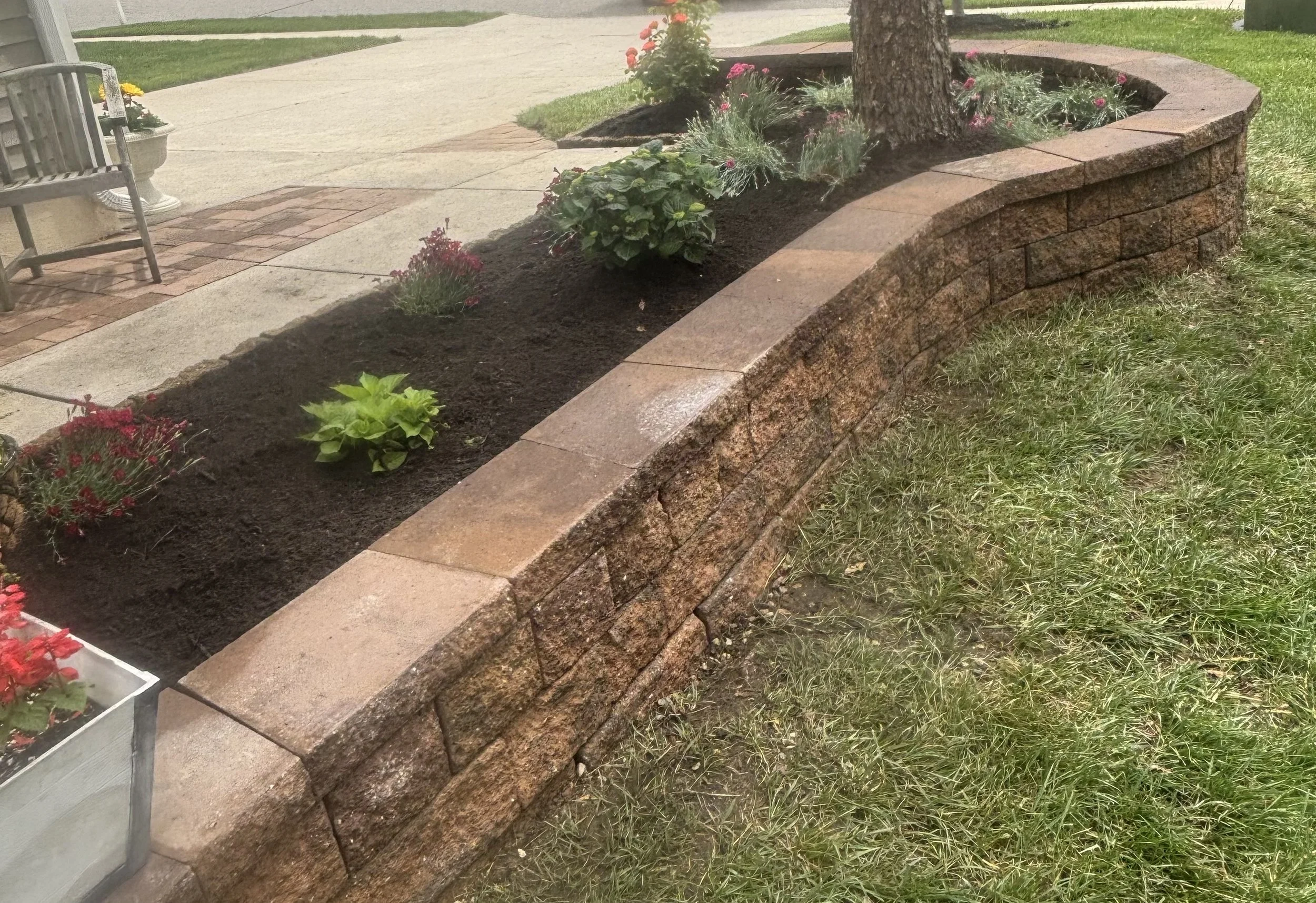 A landscaped garden bed with flowers and plants, bordered by a curved brick and stone retaining wall, and features a tree in the center. There is a concrete sidewalk and grassy area in the background.