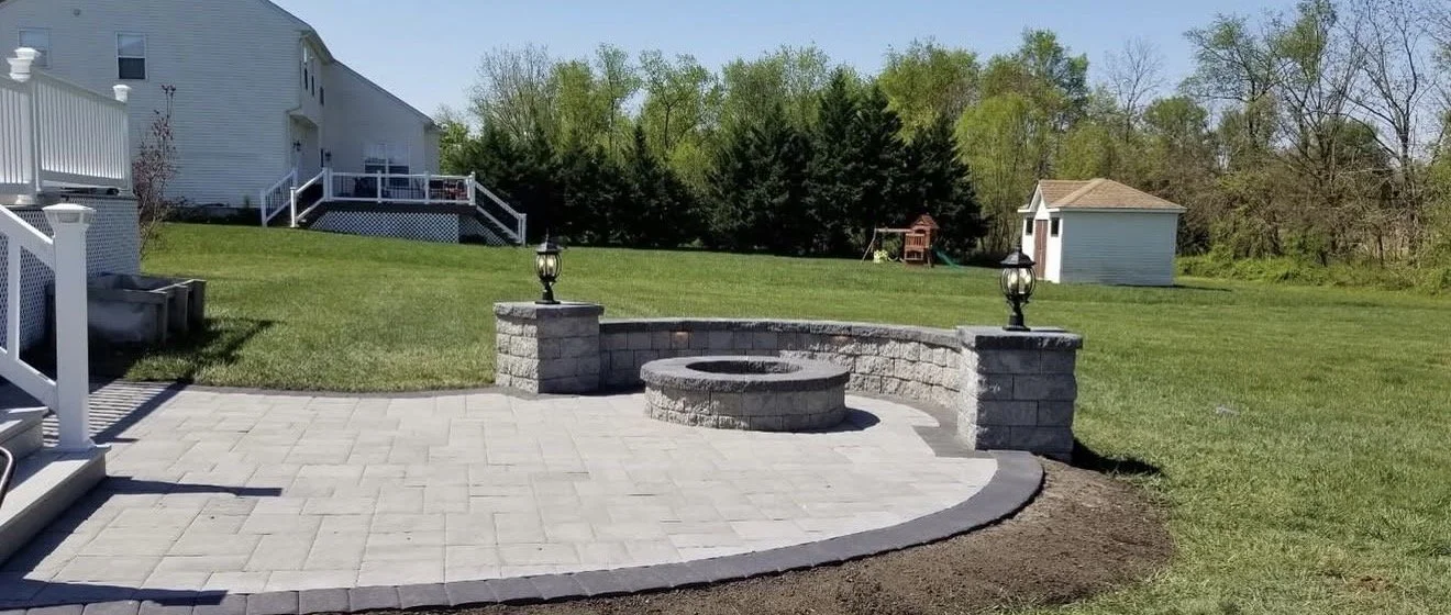 Petrol outdoor patio with a fire pit, stone wall with lamps, brick paving, and a backyard with grass, trees, and a small shed.