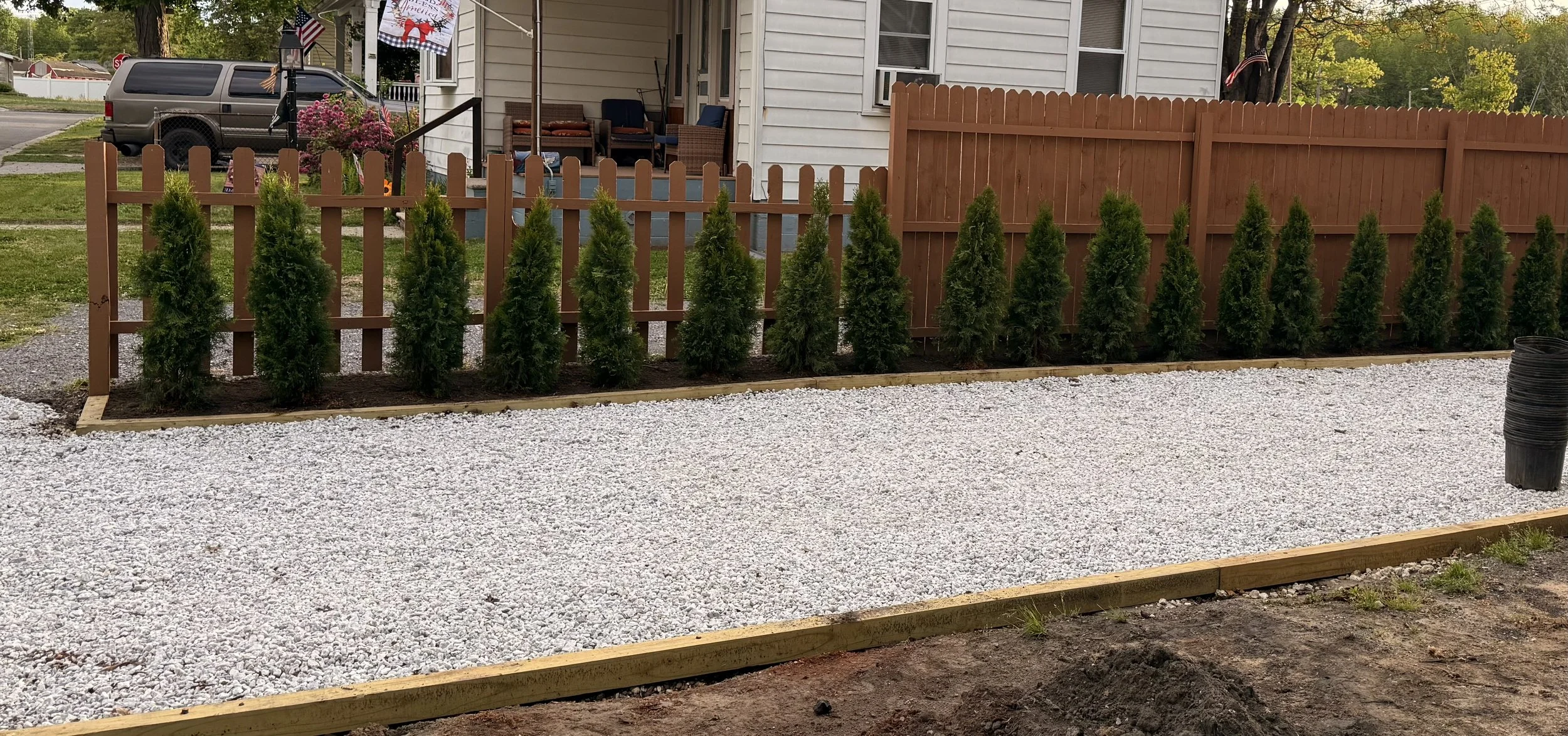 A backyard with a new wooden fence, a row of evergreen bushes planted along the fence, a gravel area bordered by wood, and a house with a porch in the background.
