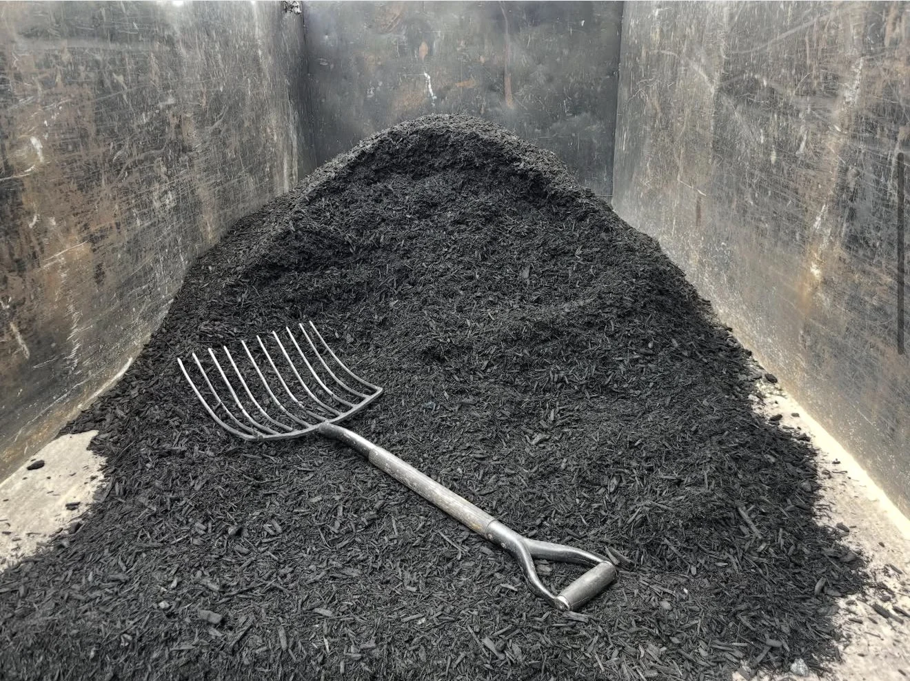 A pile of black charcoal or ashes with a metal rake lying on top, inside a metal container or wood-burning stove.