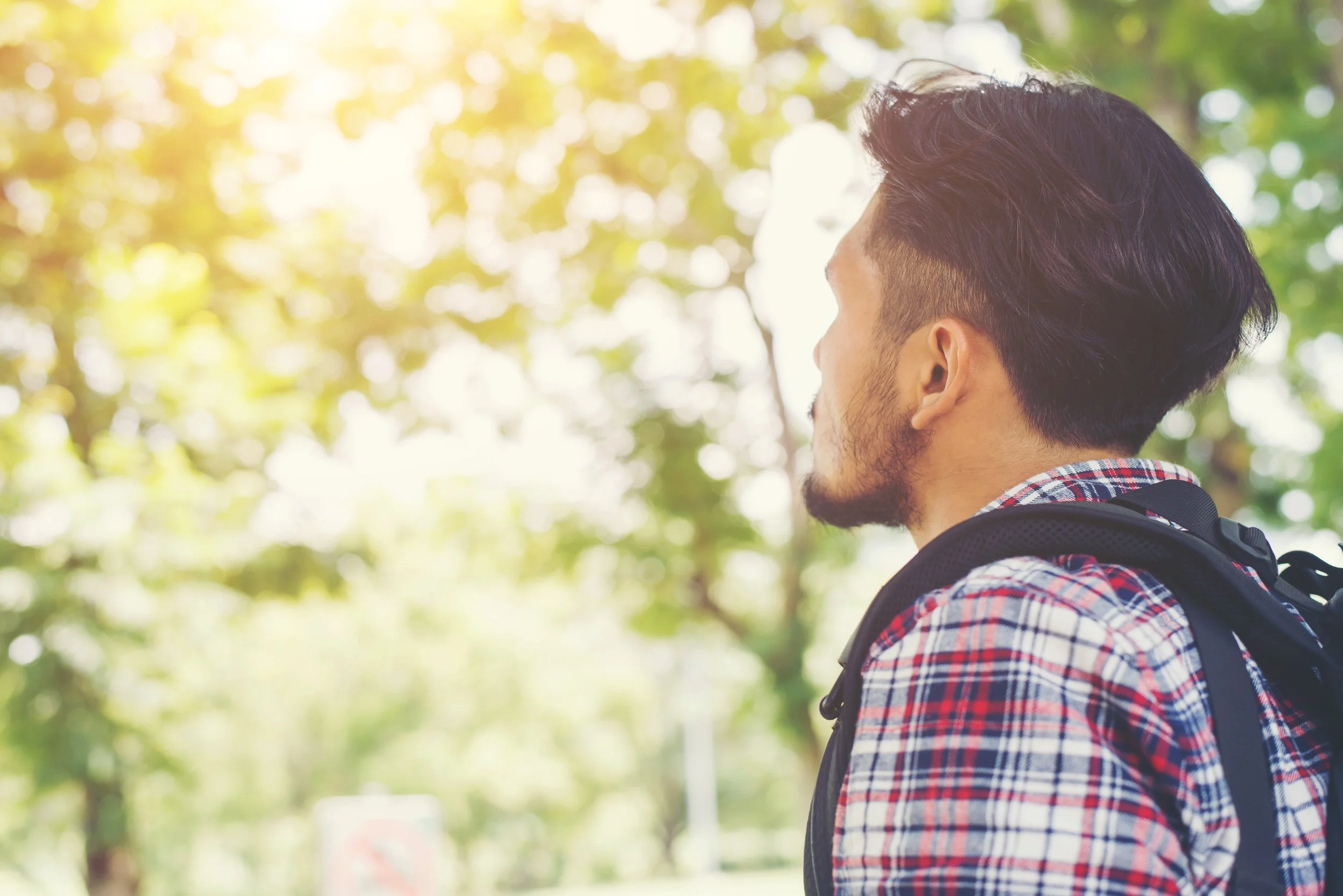 Side view of a young man with dark hair and beard, wearing a plaid shirt and carrying a backpack, outdoors with blurred green and yellow foliage in the background.