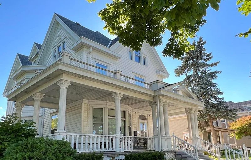 A large, white, three-story Victorian-style house with a wraparound porch supported by columns, surrounded by greenery and trees, under a clear blue sky.