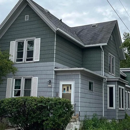 A two-story house with green siding, white trim, and a gable roof, surrounded by greenery.