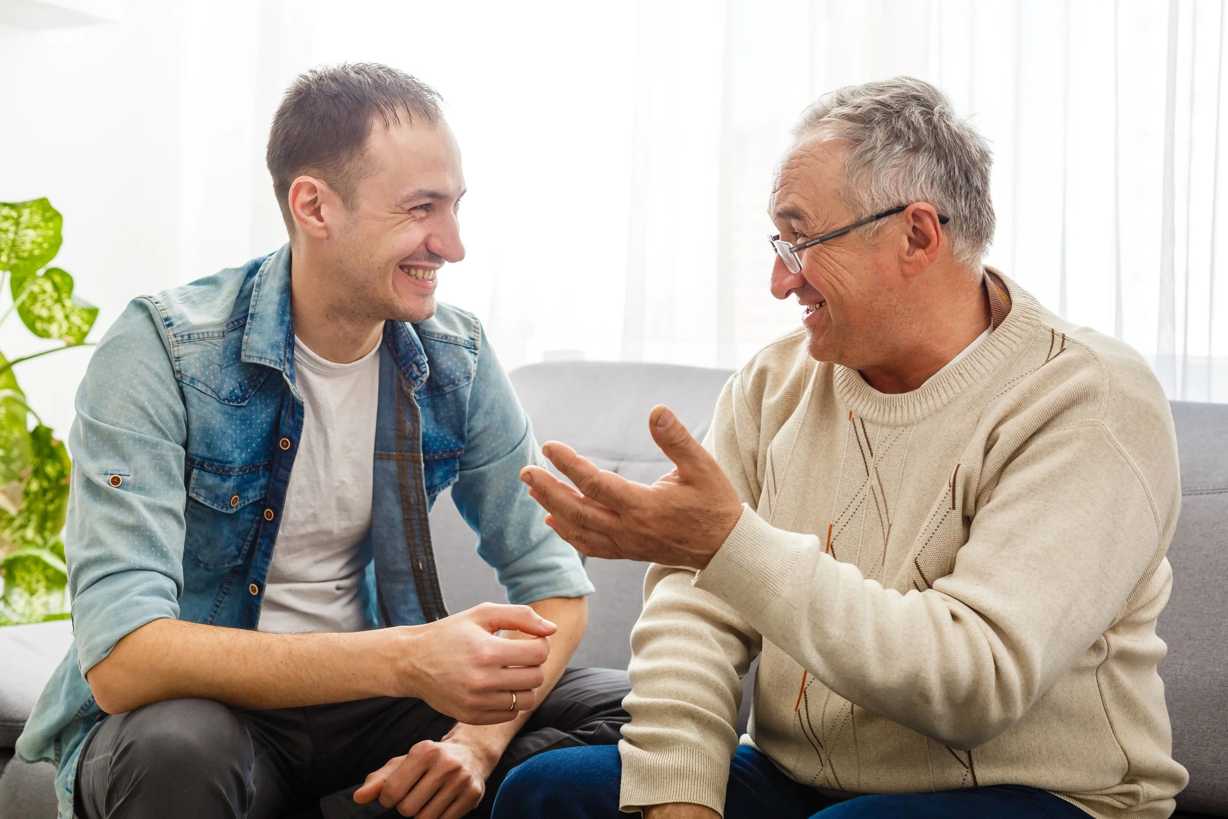 Two men sitting closely and engaging in a friendly conversation, smiling and making eye contact in a bright, indoor setting.