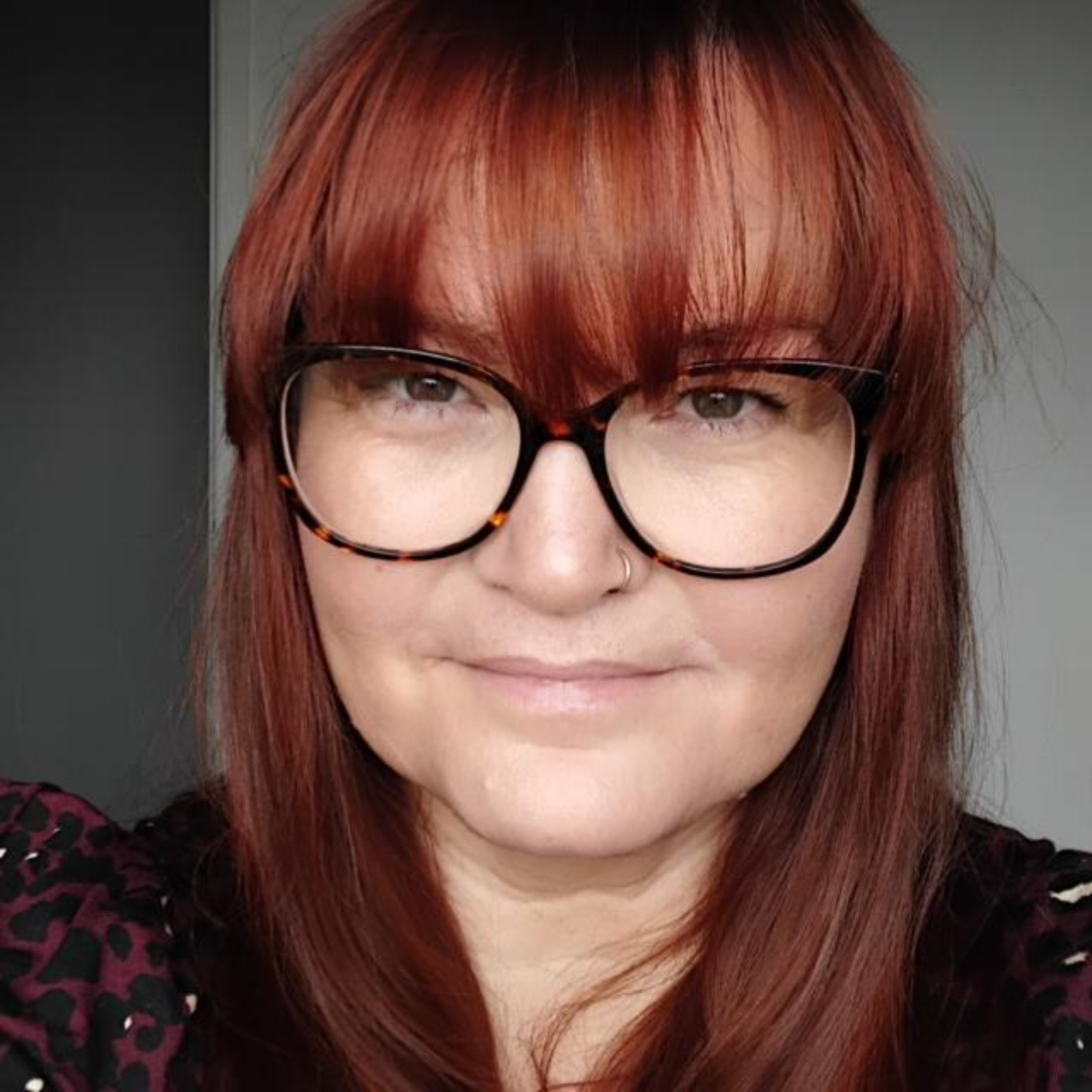 A close-up of a woman with red hair and black glasses, wearing a black shirt with pink leopard print on the sleeves.