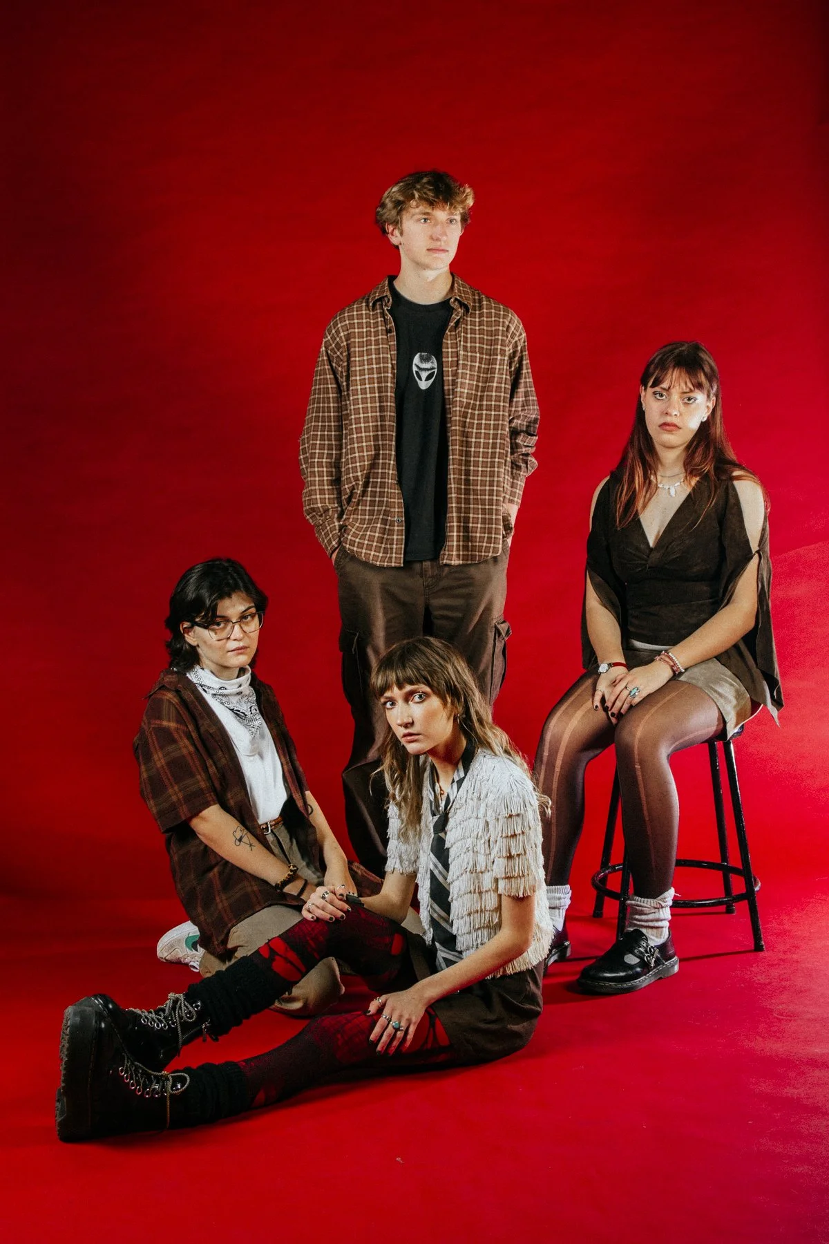 Group of four young women, one standing, three sitting, against a red background, dressed in casual and alternative fashion.