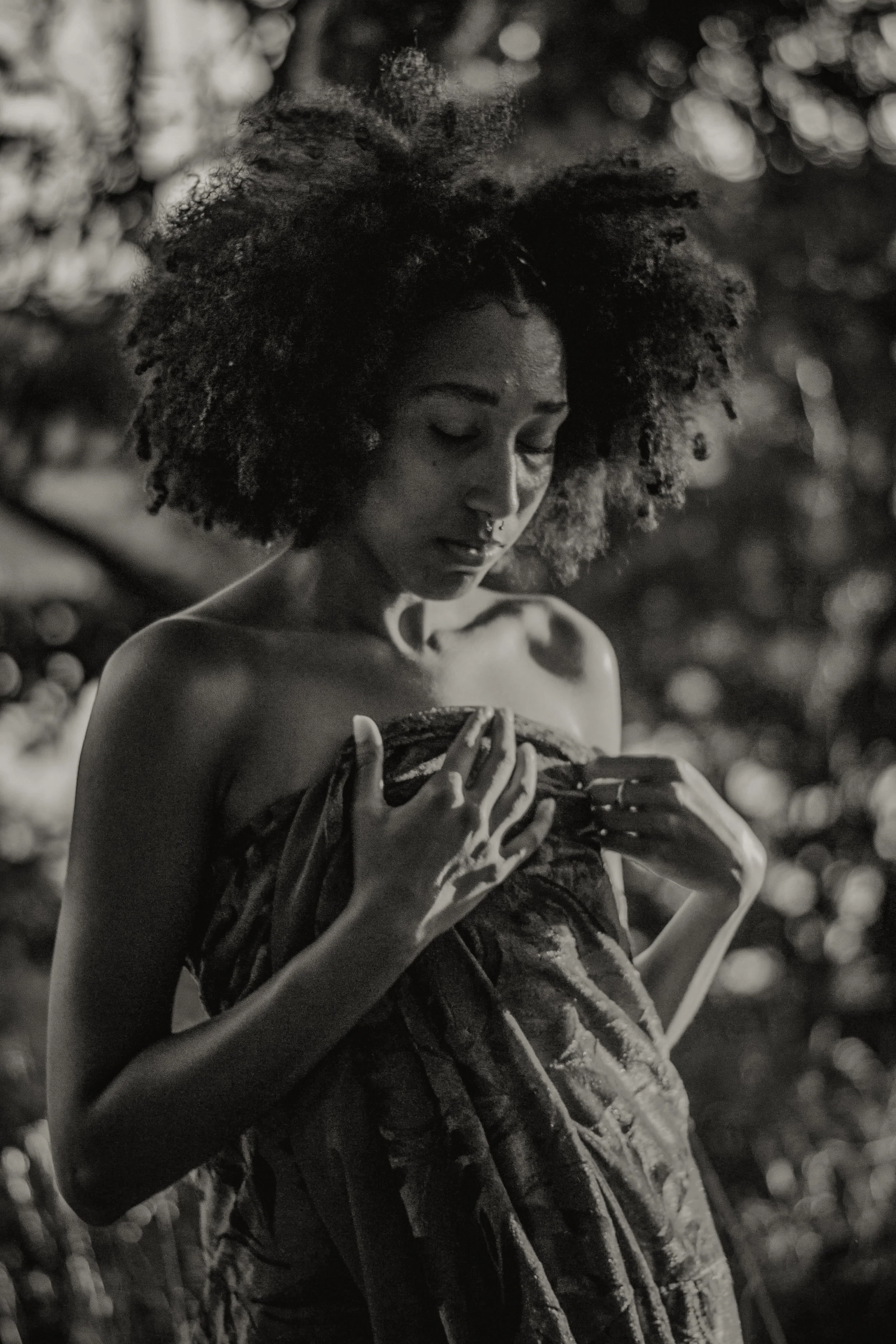 A black-and-white image of a woman with natural afro hair, looking down and holding a piece of fabric or clothing with both hands. The background is blurred with circular bokeh lights.