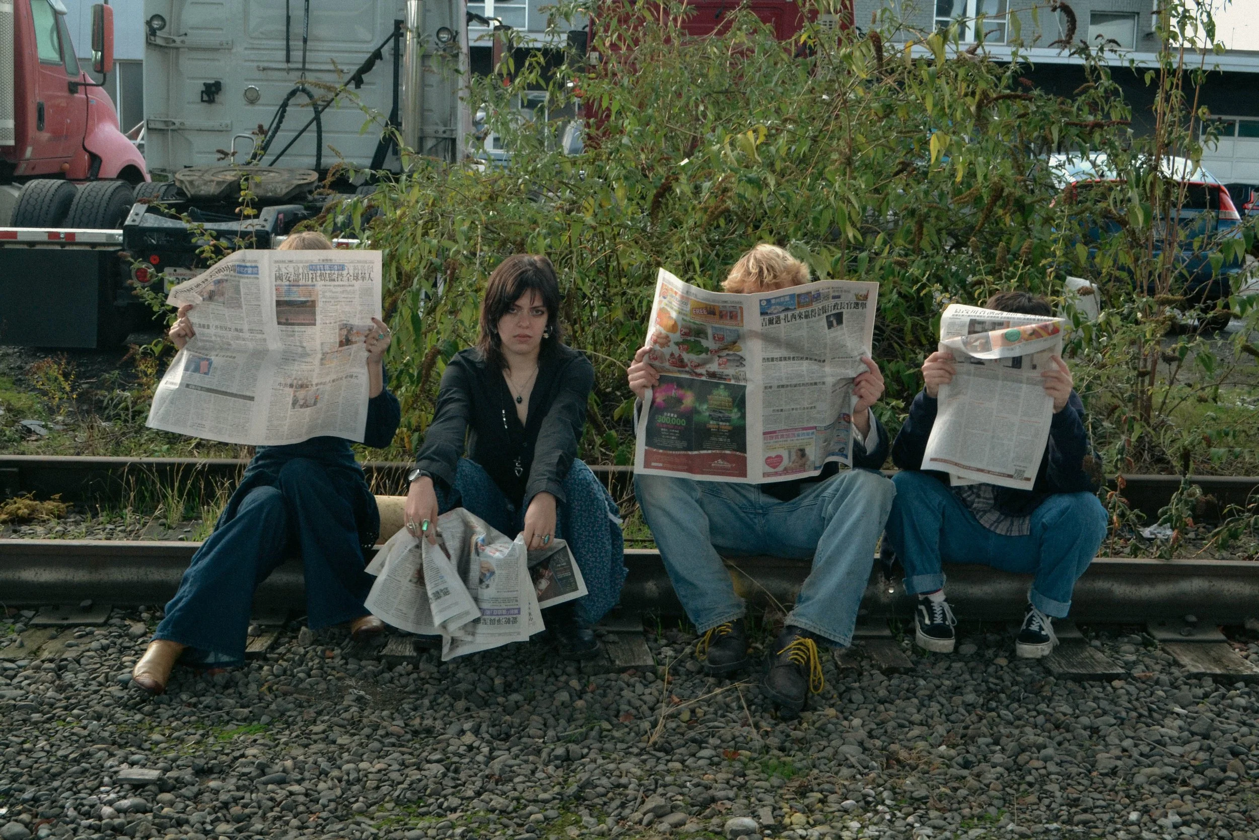 Four people sitting on a railroad track, reading newspapers, with greenery and vehicles in the background.