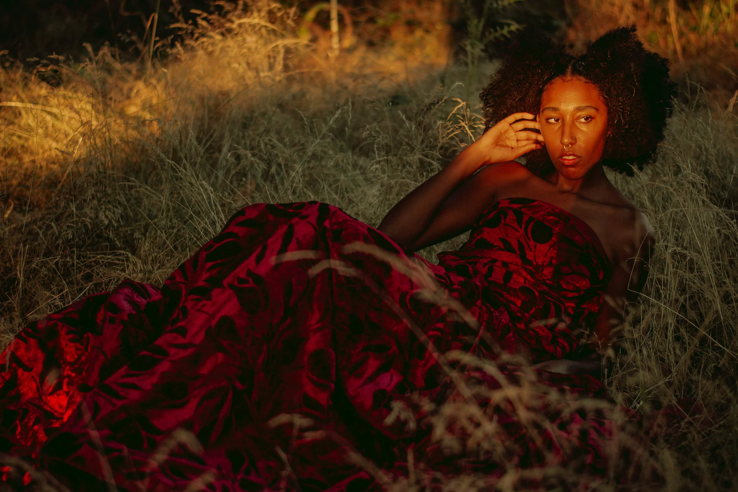 A woman with dark skin and voluminous curly hair lying on the grass, wearing a red and black patterned dress, in a natural outdoor setting during sunset.