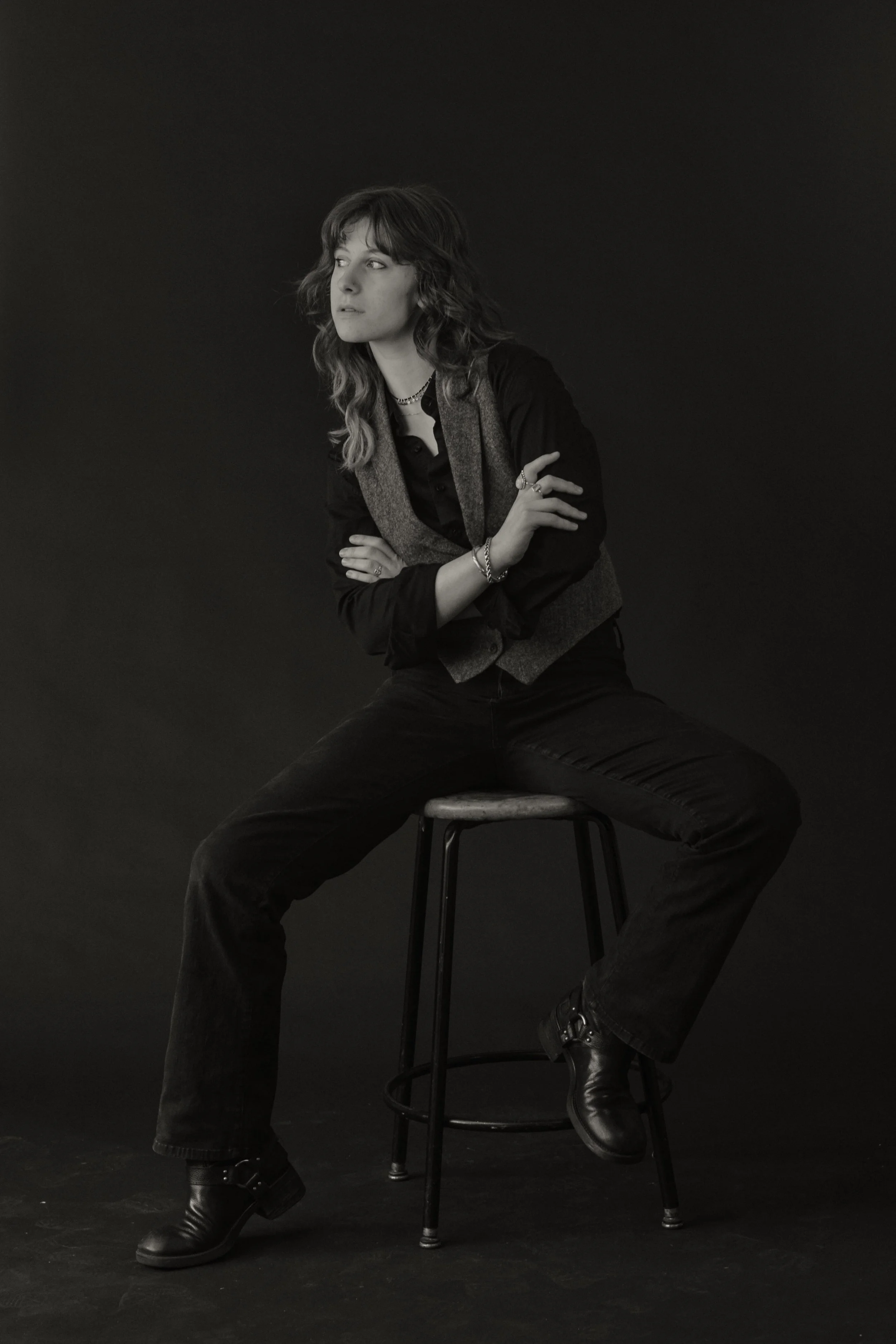 Black and white photo of a woman sitting on a stool with her arms crossed, looking to the side, wearing black pants, a dark shirt, a vest, and boots, against a plain dark background.