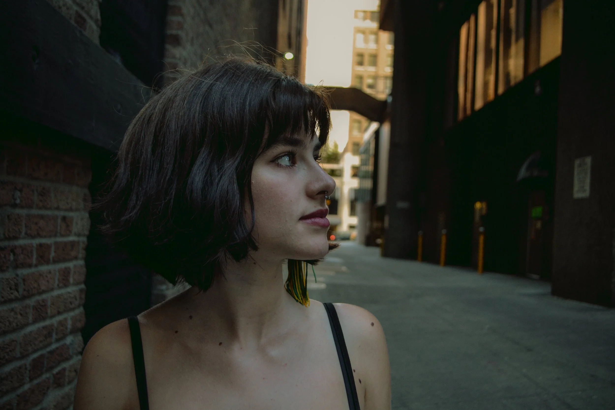 A young woman with dark, wavy hair and a septum piercing standing against a brick wall in an urban alleyway during daylight.