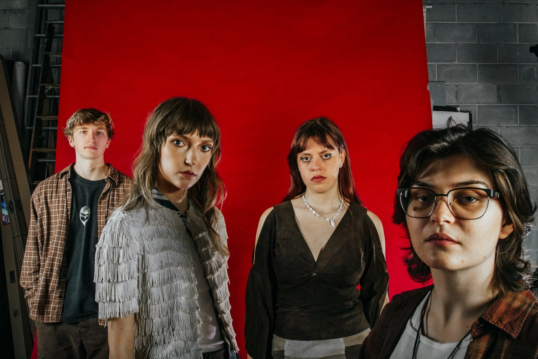 Group of four young people posing against a red backdrop, with industrial-style gray brick wall visible on the right.