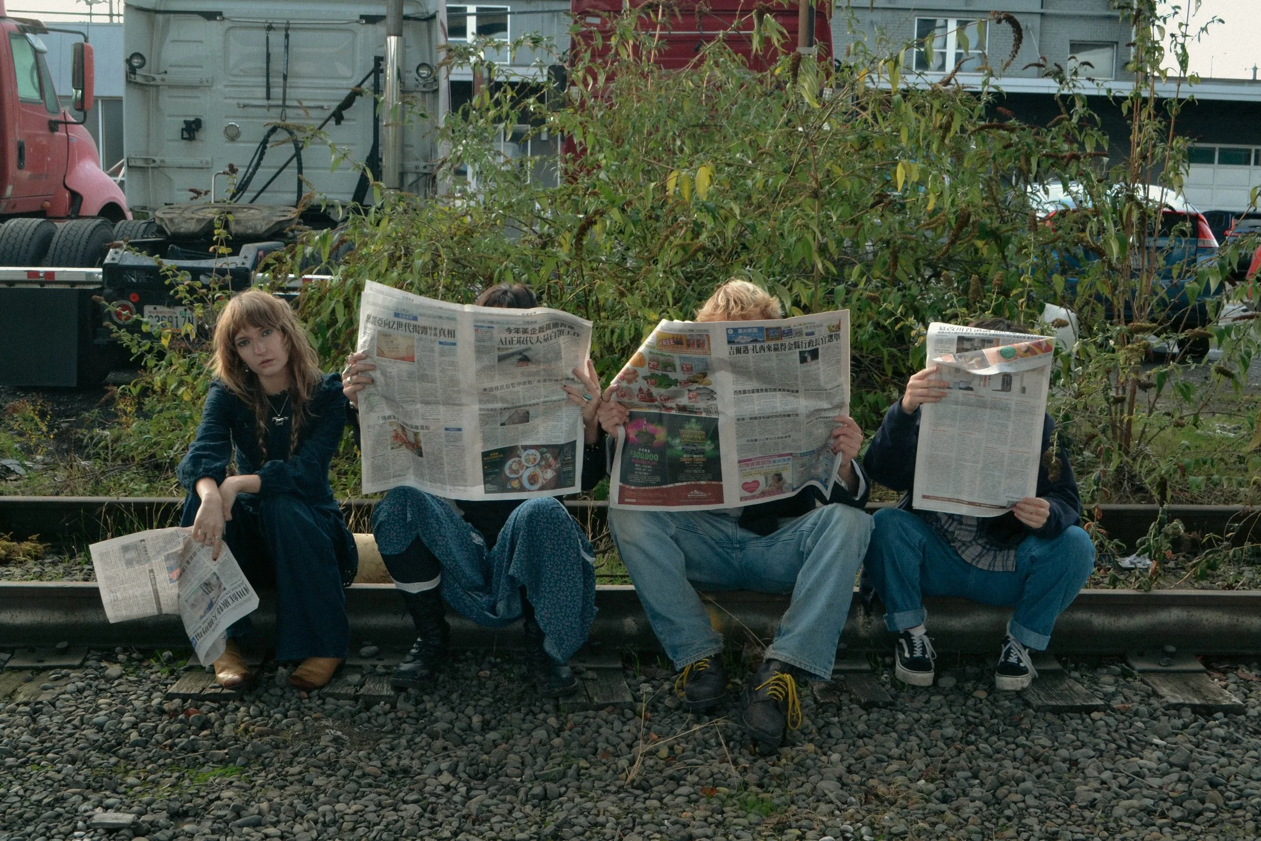 Four young people sitting on a bench next to railroad tracks, reading newspapers. One woman is looking at the camera, while the other three are focused on their newspapers. Behind them is greenery and parked cars.