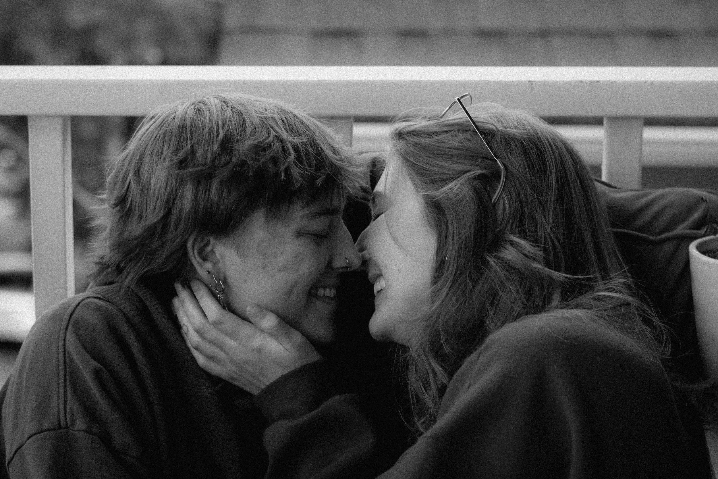 Two people are sharing an intimate moment, touching foreheads and smiling, with one person wearing glasses and the other with short hair, behind a white railing.