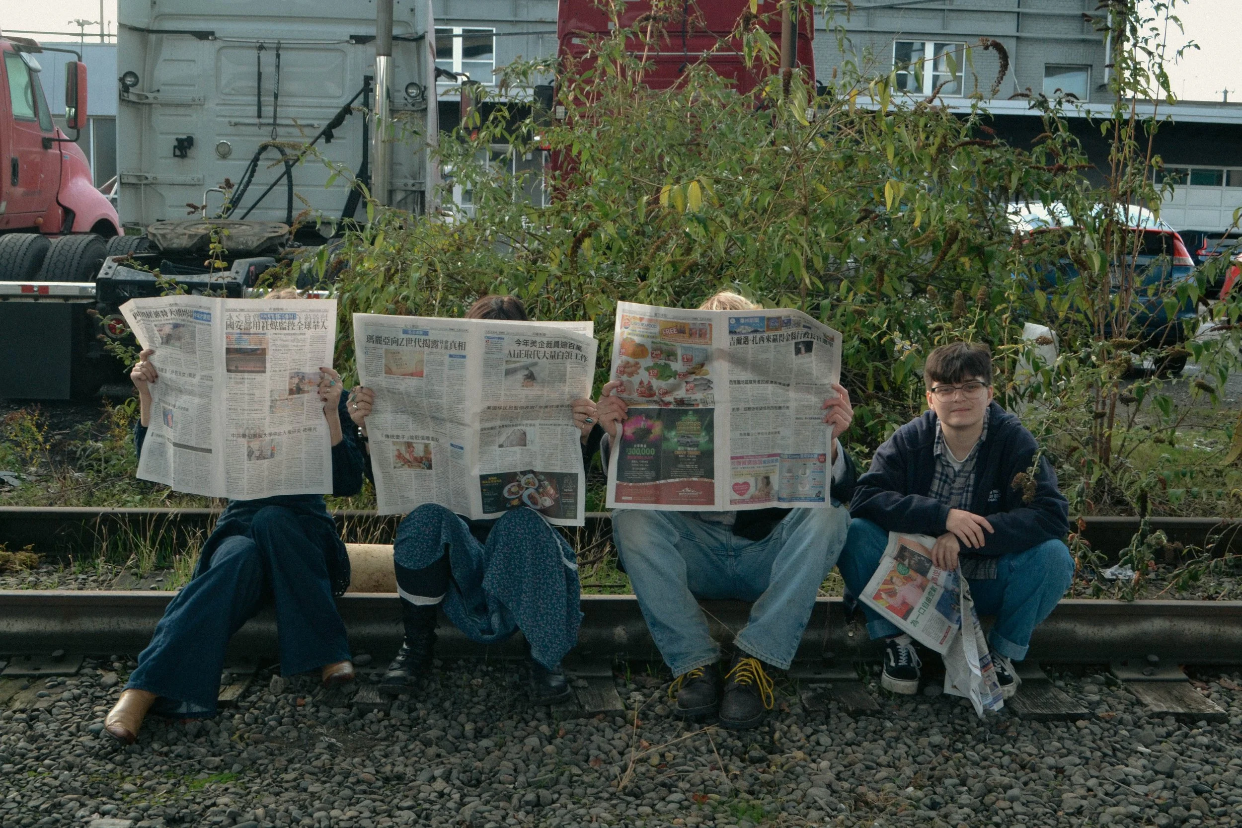 Four people sitting on a train track holding newspapers in front of their faces, with a background of trees, a red truck, and parked cars.