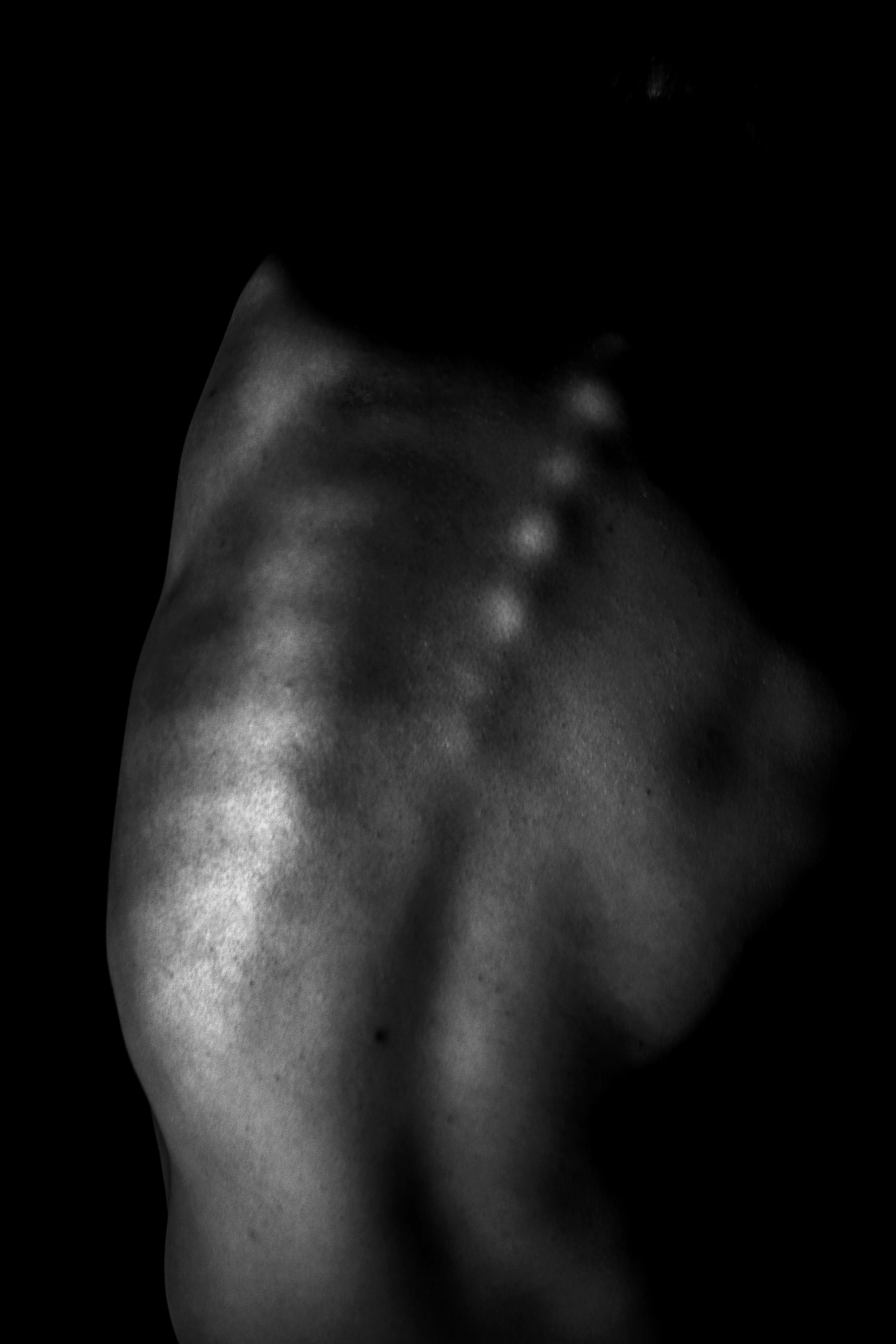 Close-up black and white photo of a person's shoulder and neck, with the person's face partially visible, shrouded in darkness.