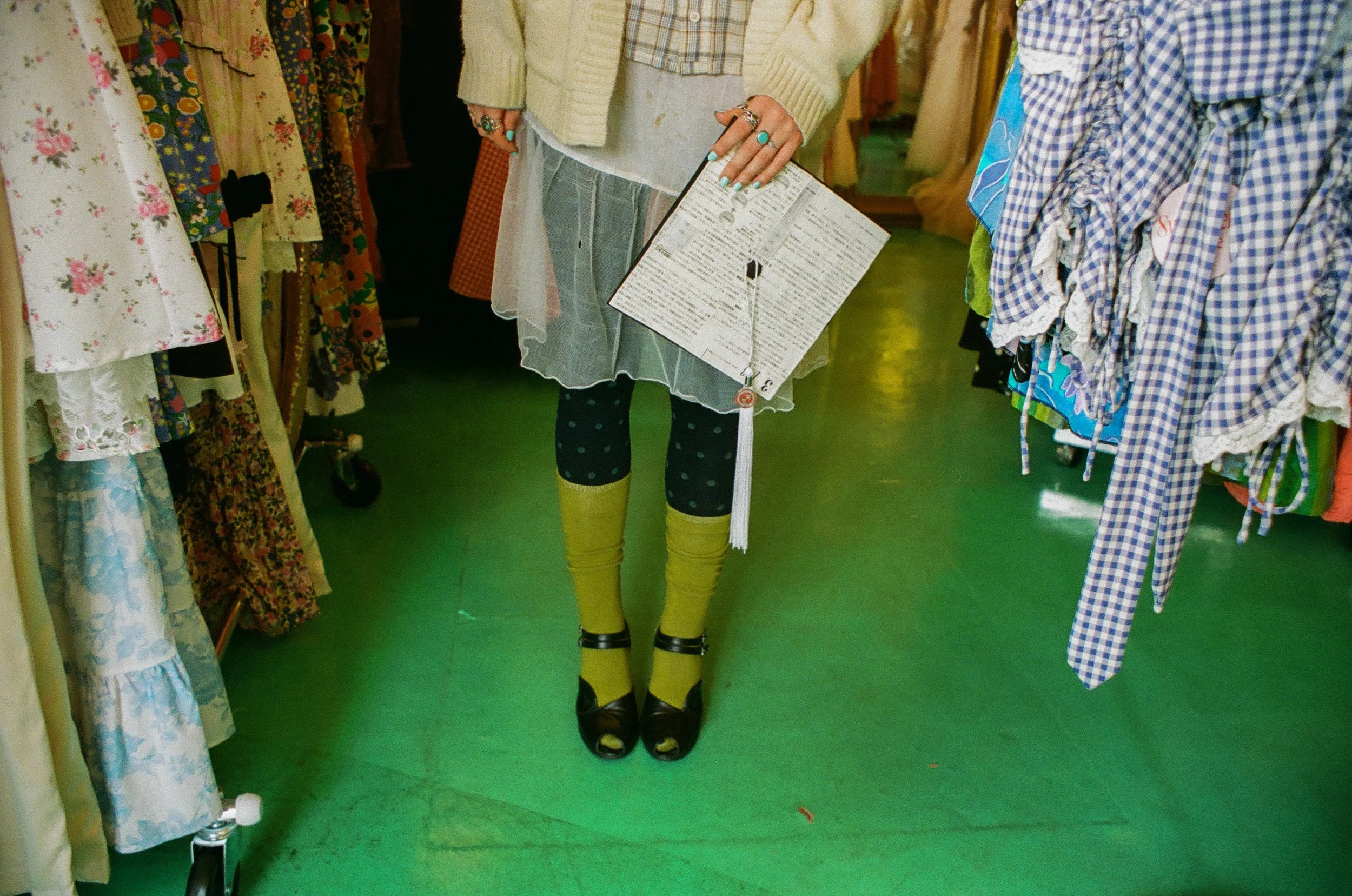 Display of a mannequin dressed in yellow leggings, black open-toe shoes, and a layered outfit including a sheer skirt and rings, surrounded by racks of colorful, patterned clothing at a thrift store.