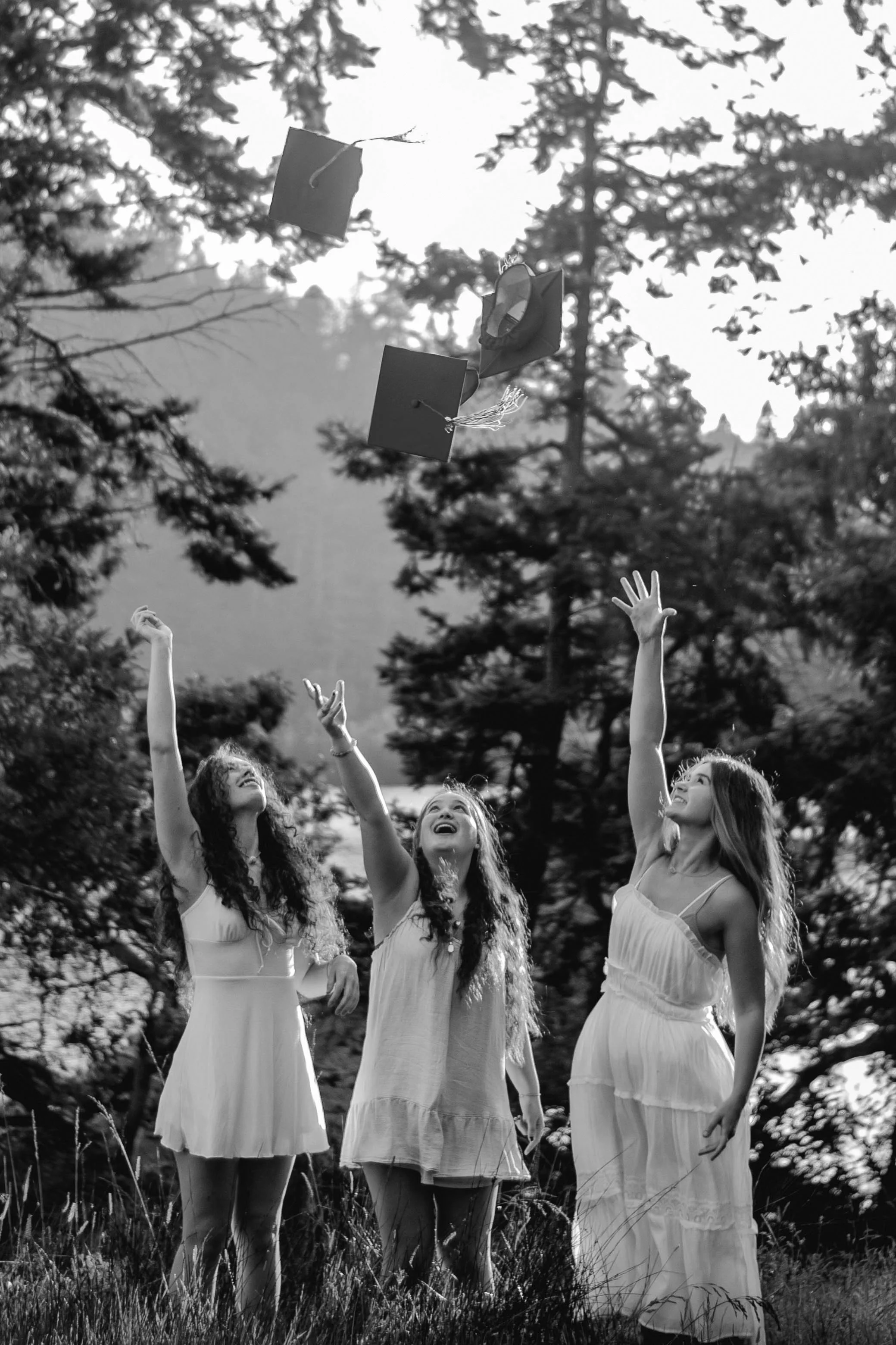 Black and white photo of three young women in white dresses outdoors, tossing graduation caps into the air, surrounded by trees and grass