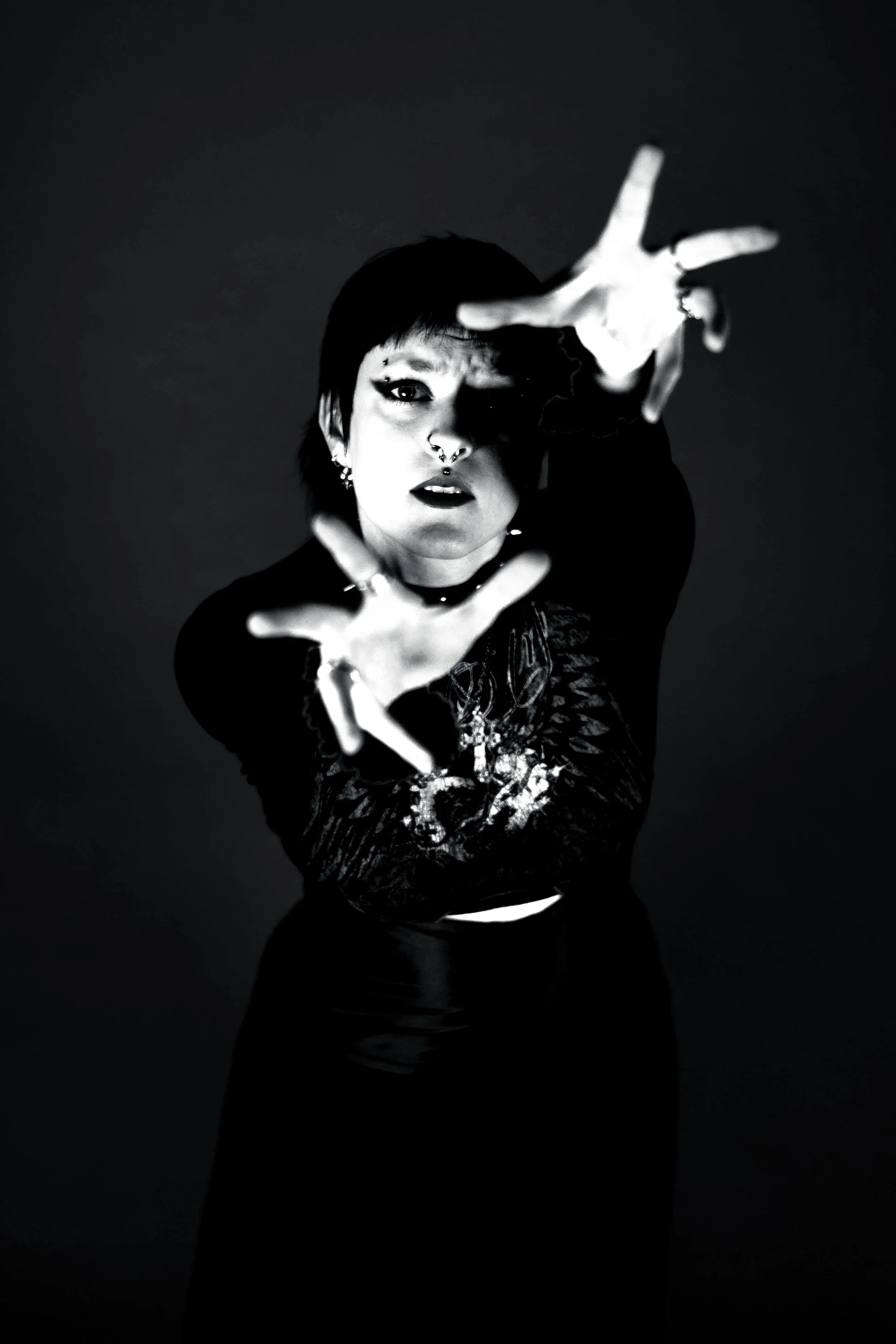 A person with dark hair and multiple facial piercings extending their hands towards the camera, creating a dramatic and intense pose in a black and white photo.