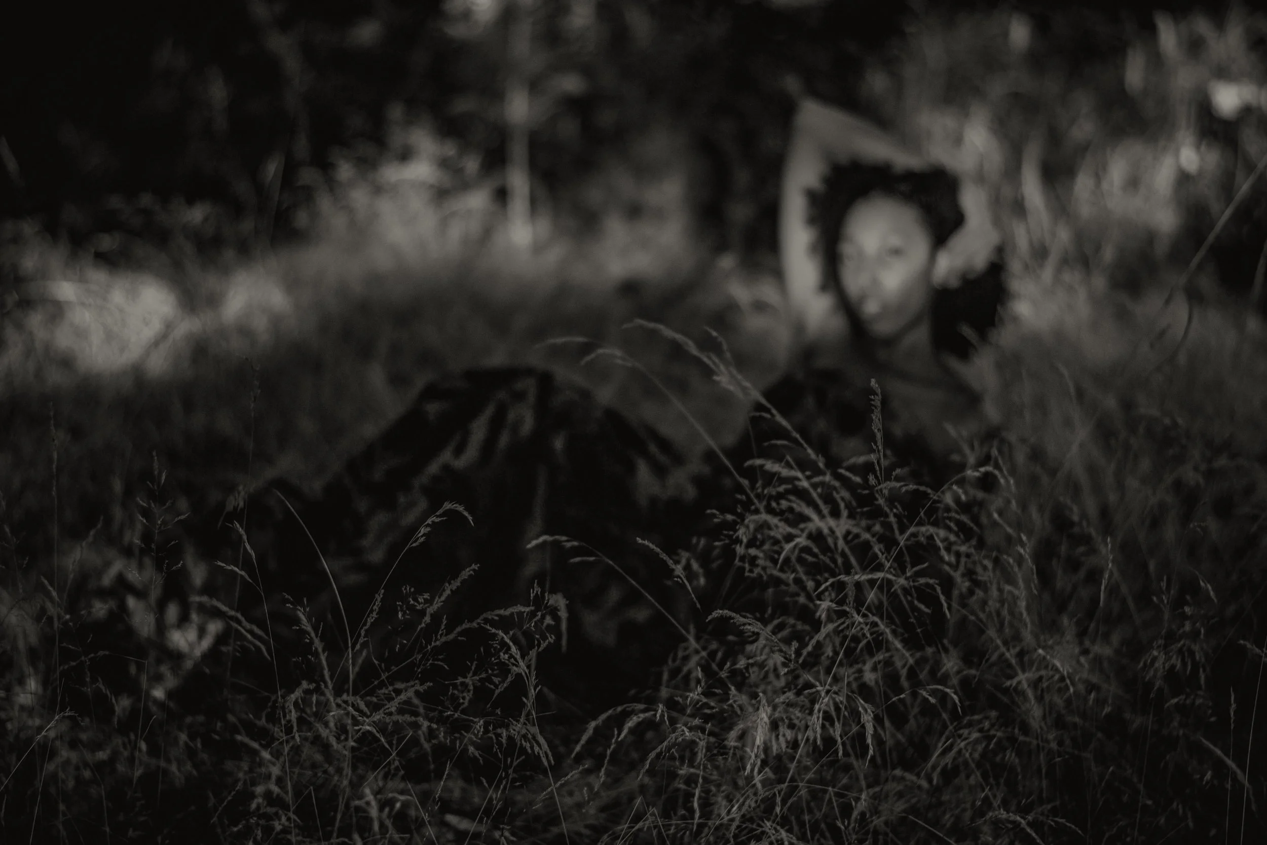 A woman with dark hair lying on the grass with a pen in her hand, surrounded by plants and greenery at night.