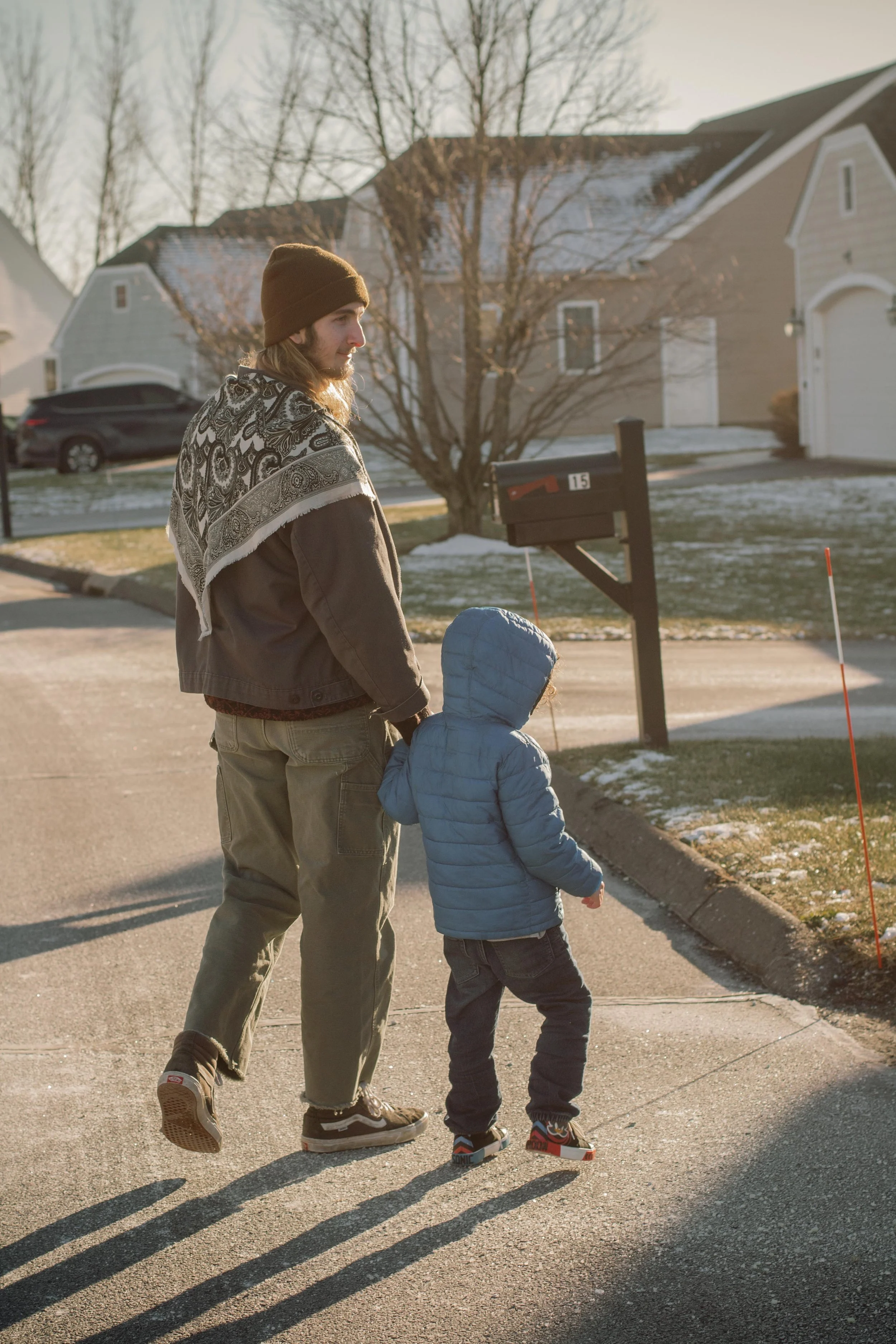 A man and a young boy walking on a suburban street during winter. The man has long hair, a beard, and is wearing a brown beanie, a patterned scarf, a brown jacket, khaki pants, and sneakers. The boy is wearing a blue puffer jacket, a knit cap, and sneakers. Shadows are cast on the pavement by the setting sun, and there is snow on the grass and rooftops.