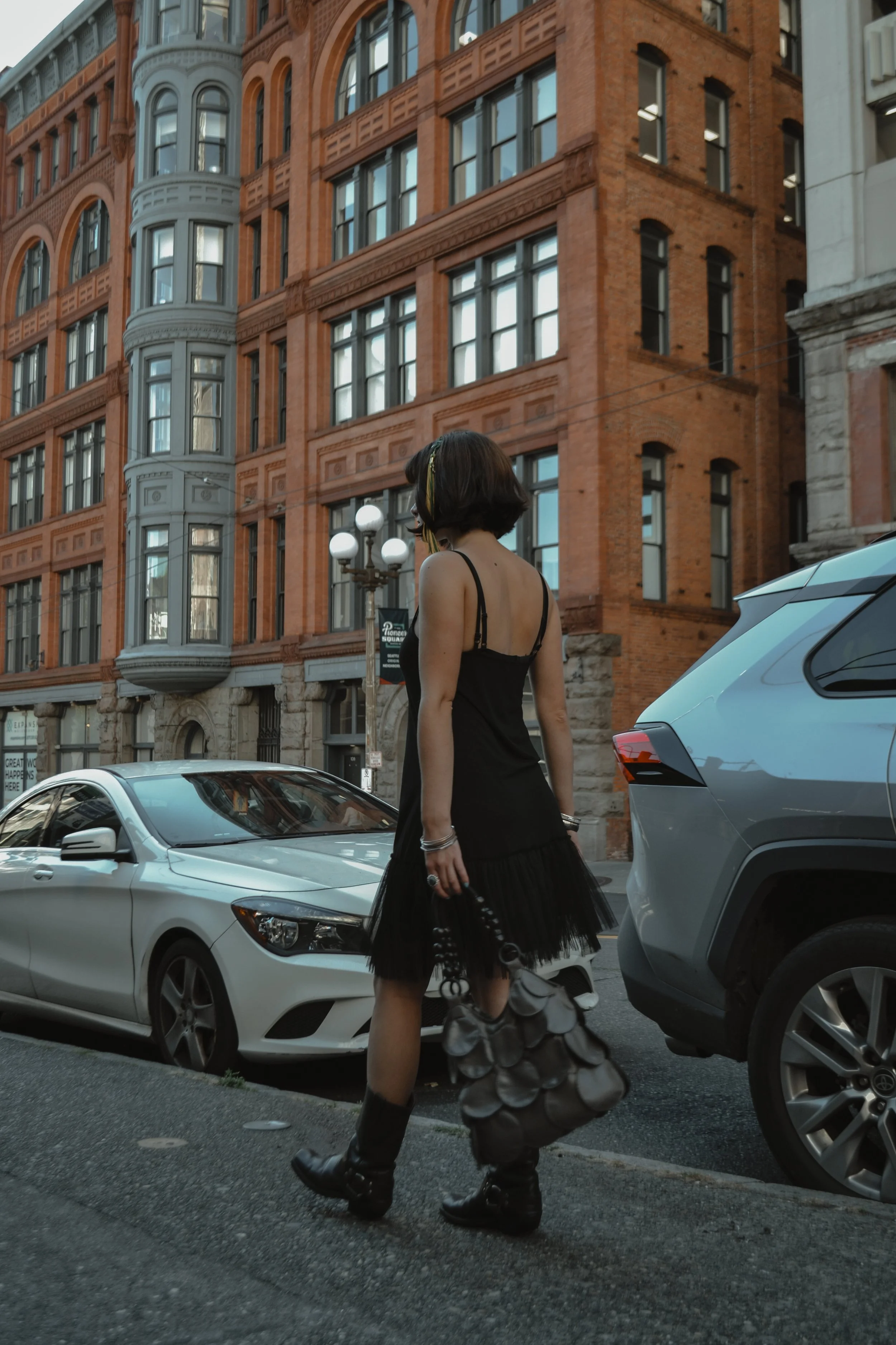 A woman with short dark hair in a black dress and black boots walking on a city street near parked cars, with historic red brick buildings in the background.