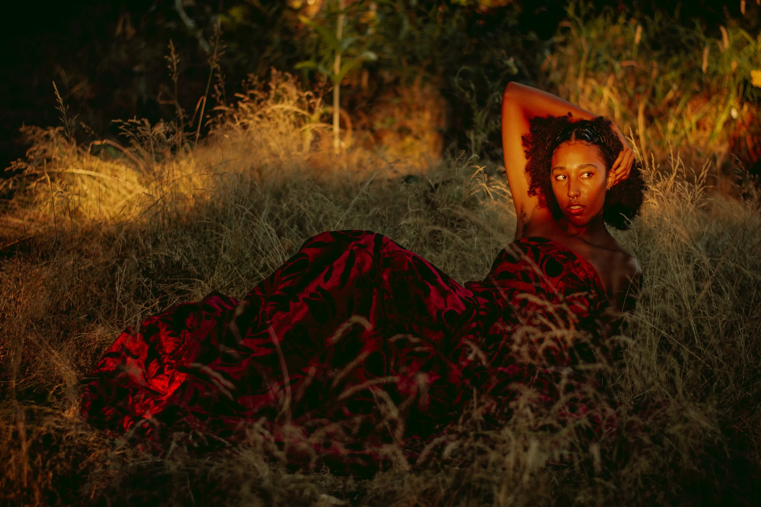 A woman with curly hair and dark skin, lounging on the ground in a field of tall grass at sunset. She is wearing a red patterned dress and has her arms raised behind her head, looking at the camera.