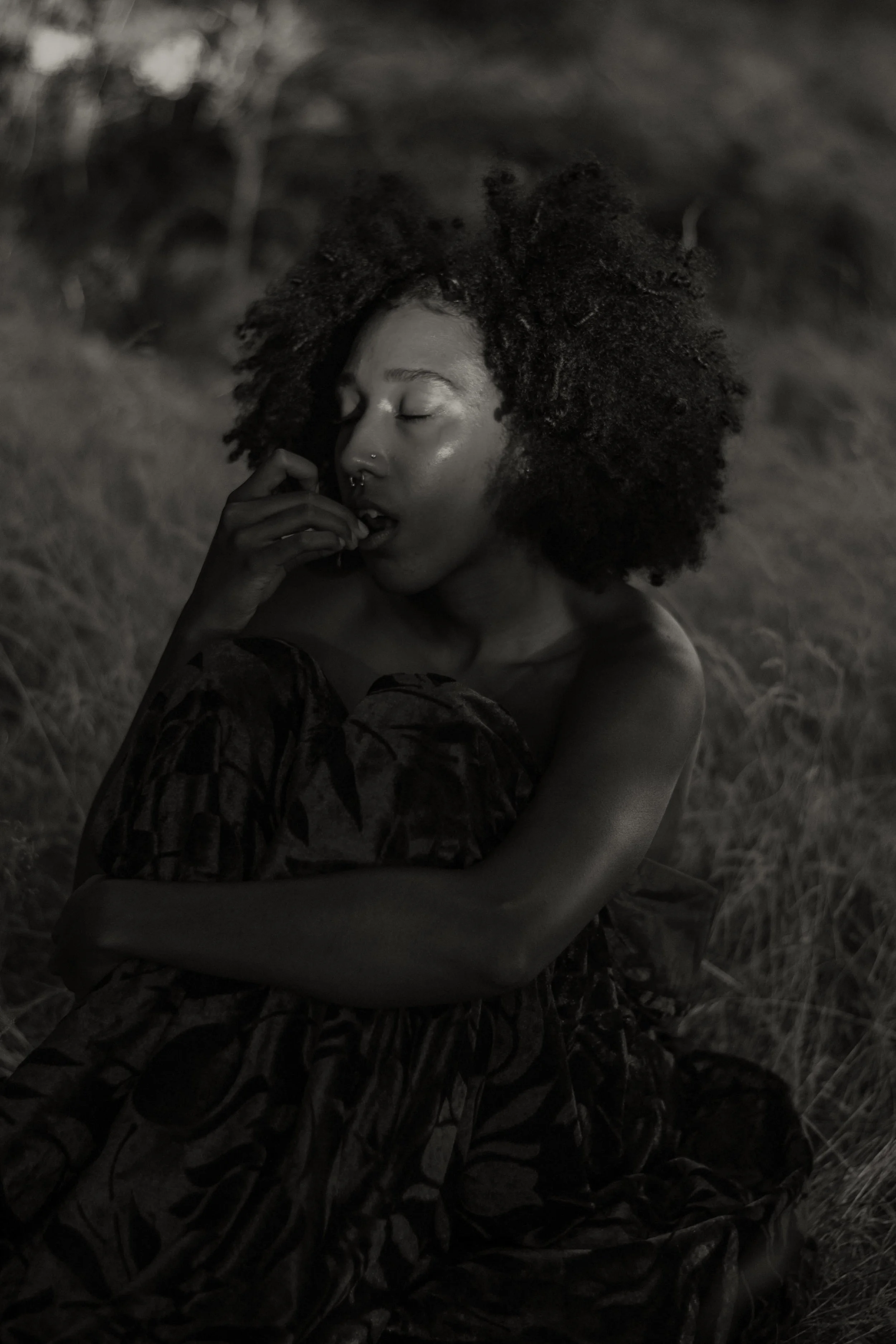 A woman with curly hair and a septum piercing, sitting outdoors in a natural setting. She has her eyes closed and hand near her face, with grass and blurred foliage in the background. The photo is in black and white.
