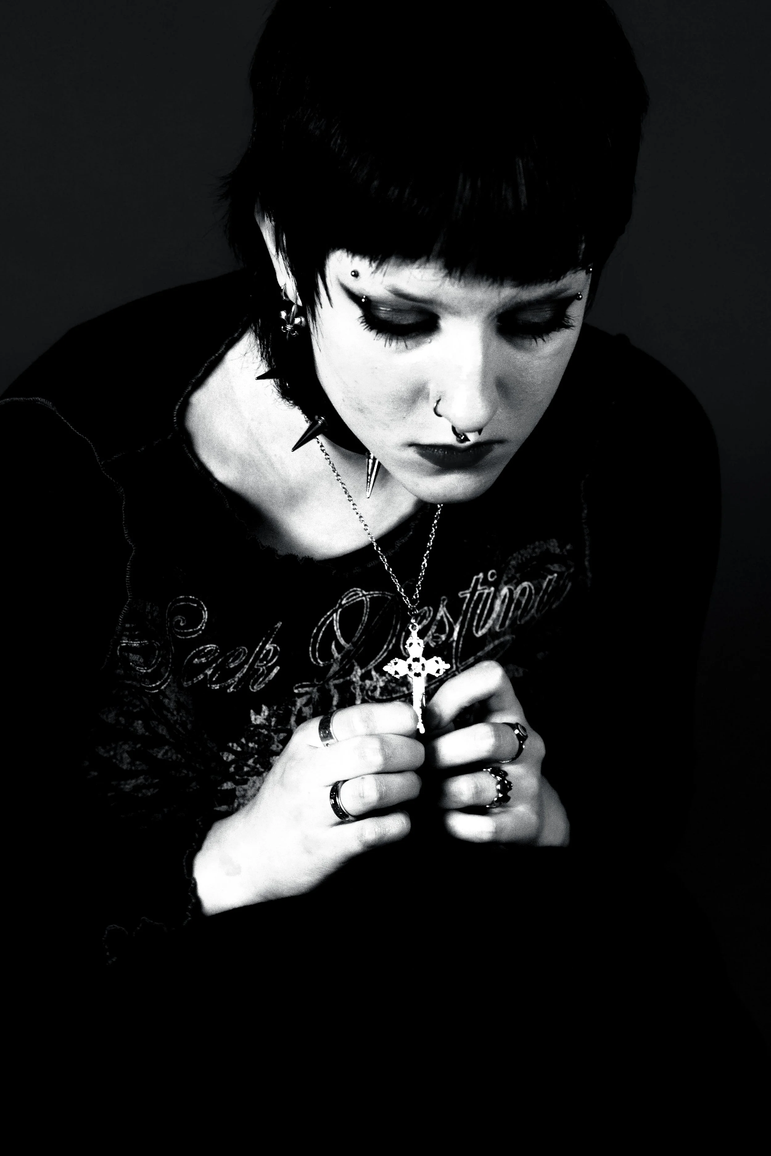 Black and white portrait of a woman with short hair and multiple earrings, wearing a spiked collar and holding a cross necklace with both hands, looking downward.
