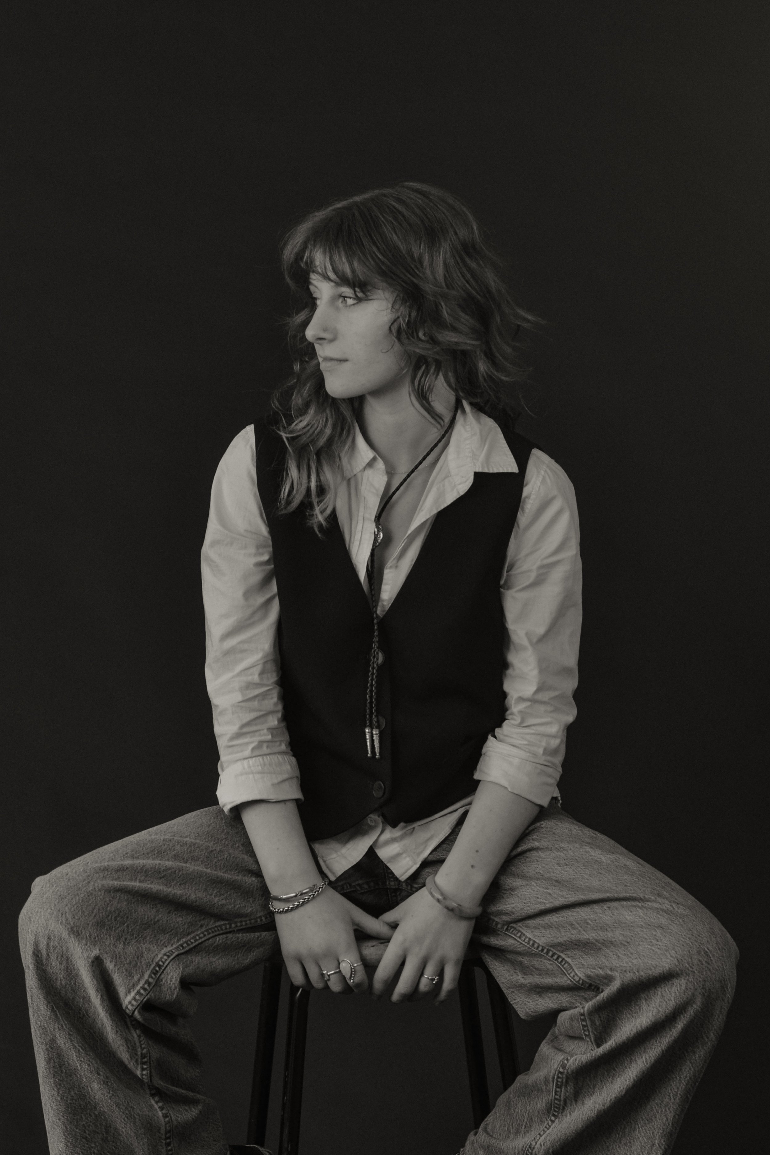 Black and white photo of a woman sitting cross-legged on a chair against a plain dark background, looking to her left.