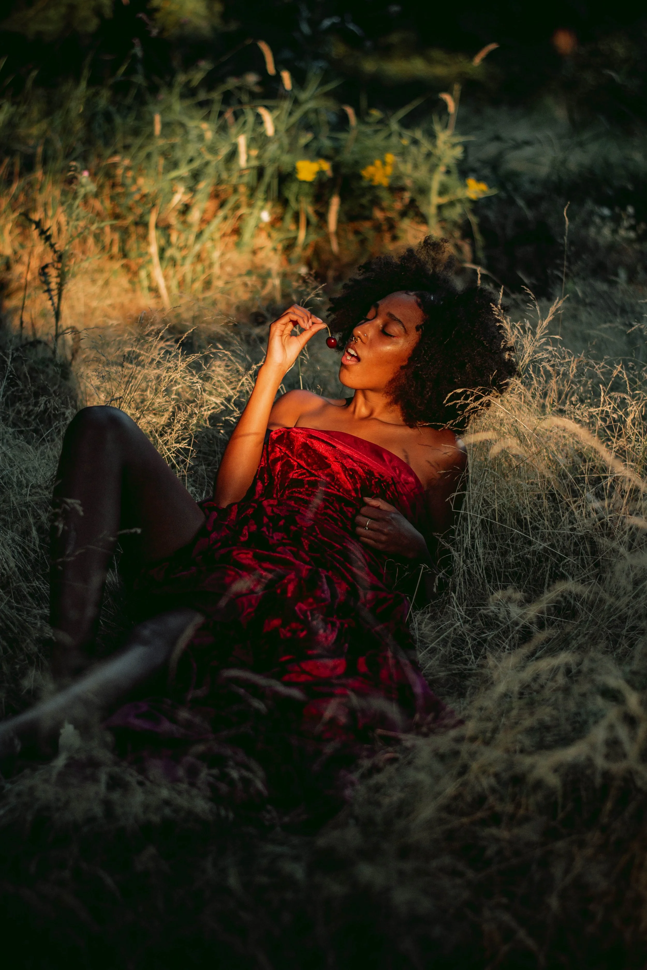 A woman with curly hair lying on the ground in a field of tall grass, holding a cherry near her mouth, dressed in a red strapless dress, during sunset or twilight.