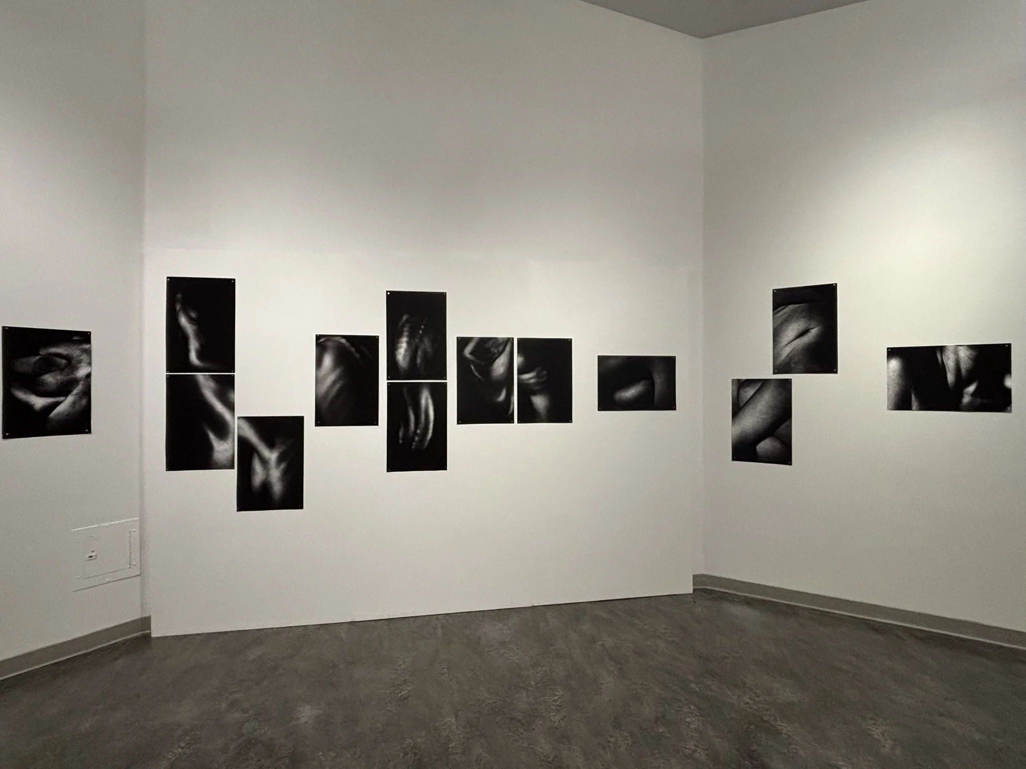 Gallery wall display with black and white photographic prints arranged in rows on white walls inside an art gallery room.