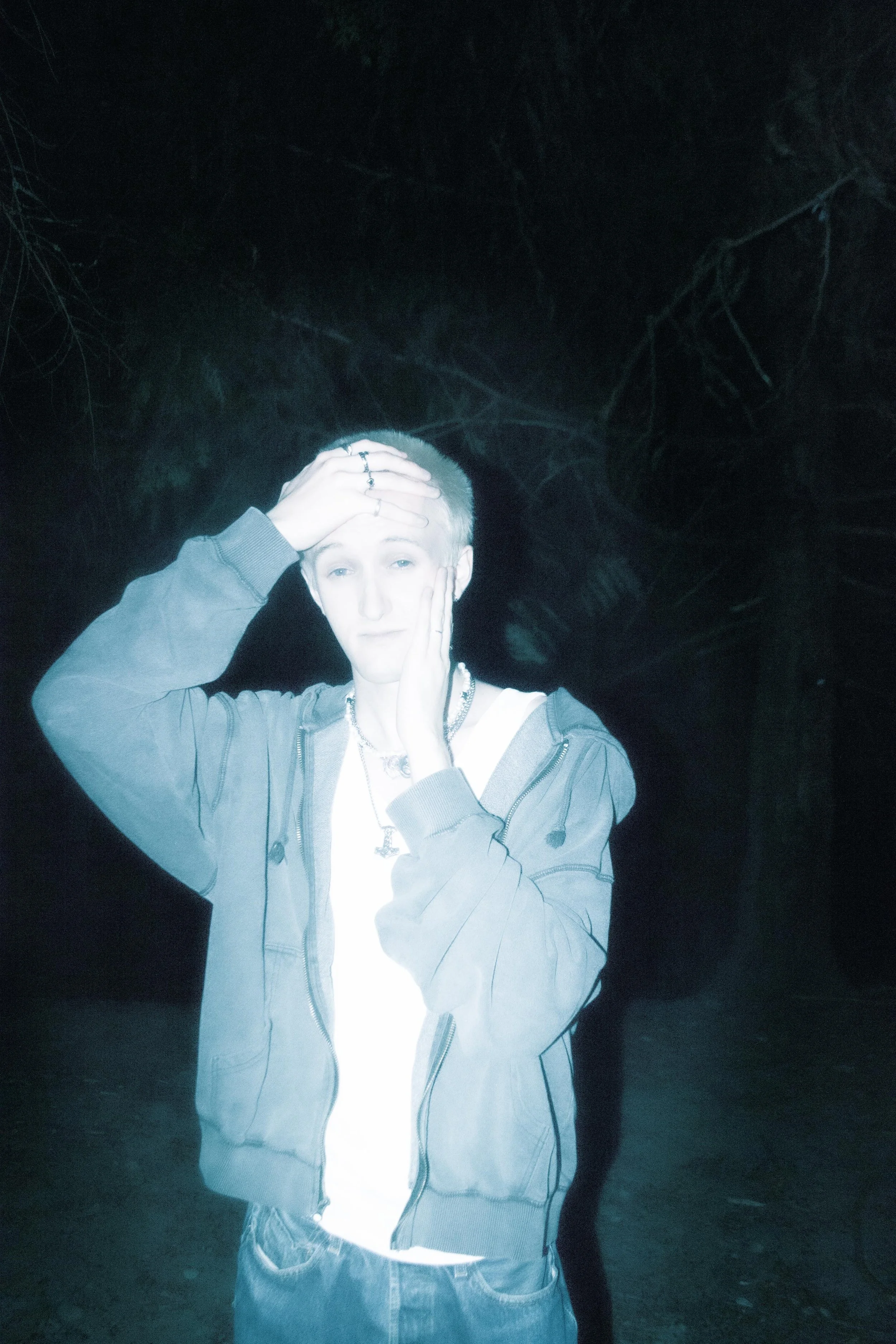 A person with short, light-colored hair, wearing a gray hoodie over a white shirt, standing outdoors at night surrounded by dark trees, holding their head with one hand and resting their face in the other hand.