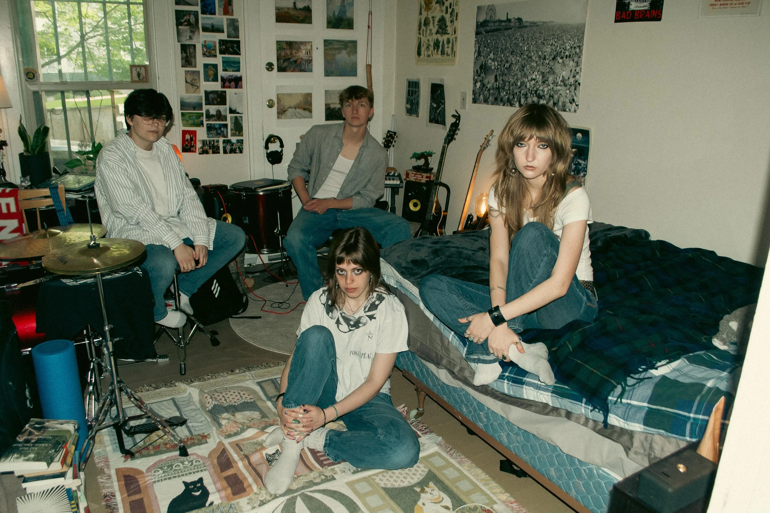 A group of five young people in a cluttered bedroom with musical instruments and posters on the wall, some sitting on a bed, one on the floor, and one sitting on a chair.