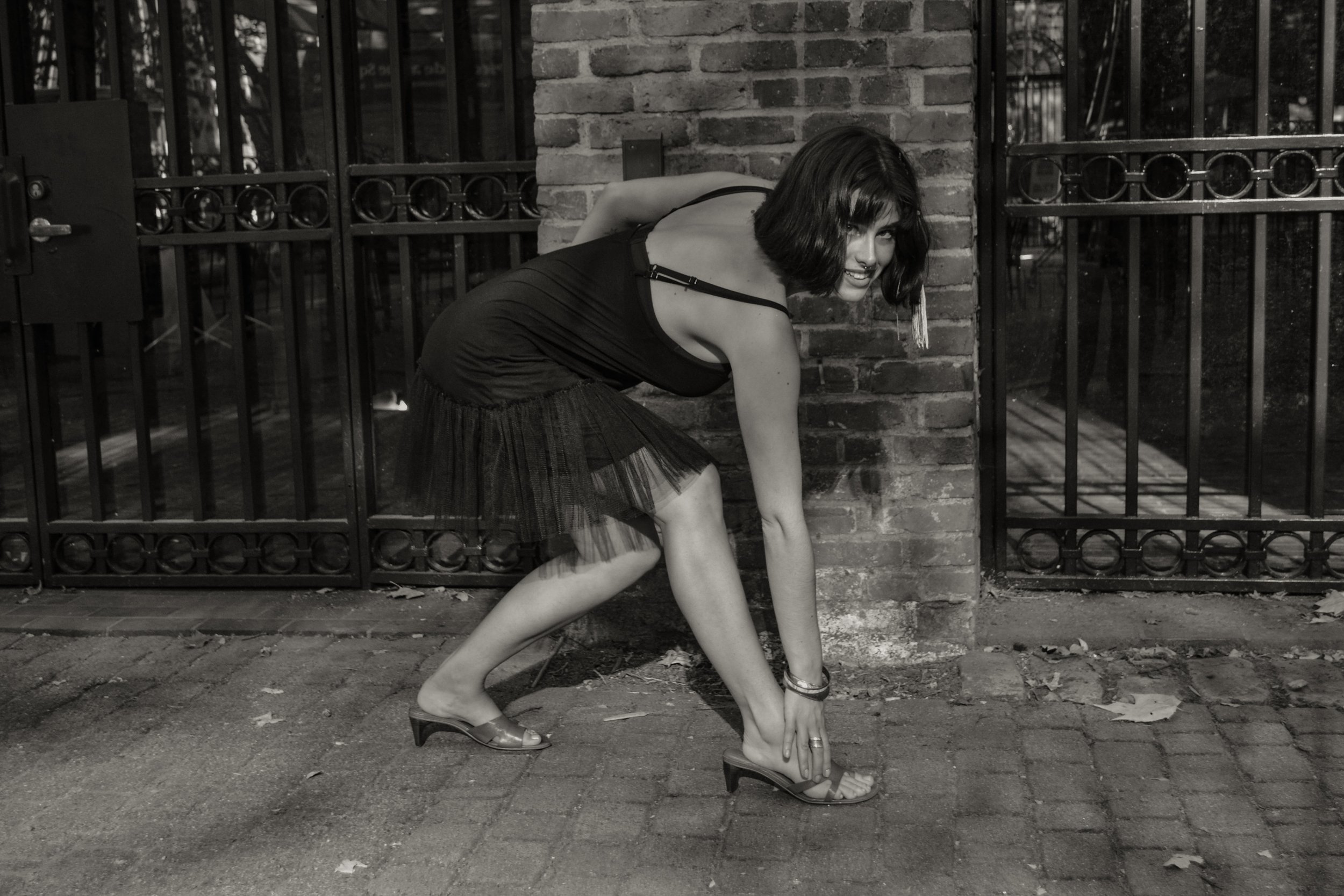 A woman with shoulder-length dark hair, wearing a black tank top, plaid skirt, and high heels, is kneeling on the ground, leaning forward with one hand touching her shoe, in front of a brick wall and metal gate.