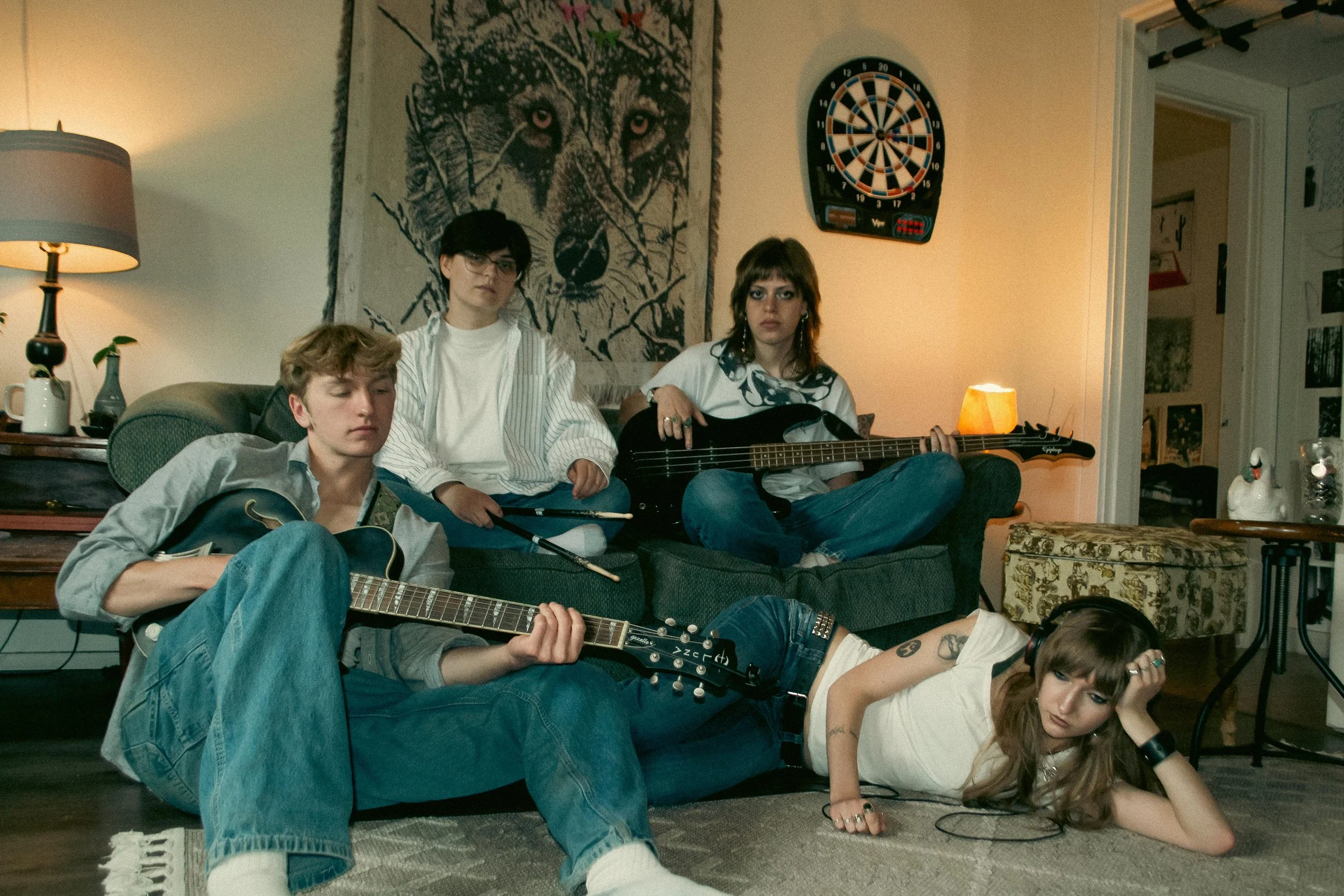 A group of young people in a living room, some sitting on the sofa and others lying on the floor, engaged in playing guitars, with a woman holding a bass guitar. There is a large wolf tapestry on the wall behind them, and a dartboard next to it. The 