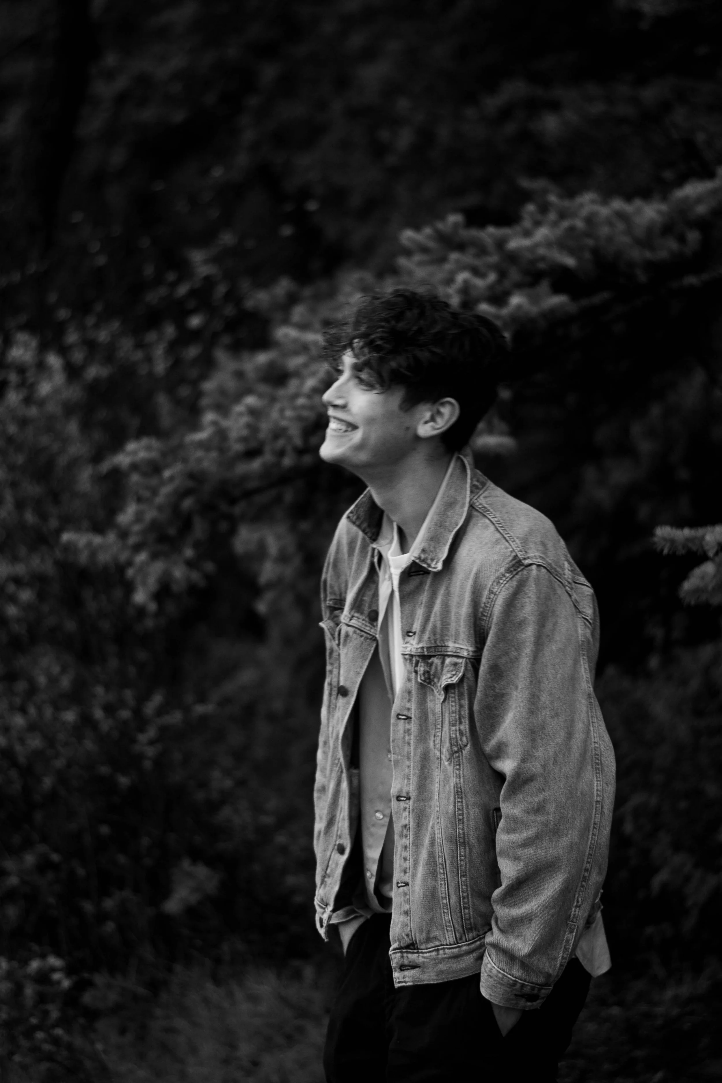 A young man with curly hair and a smile, wearing a denim jacket, walking outdoors near trees. Black and white photography.