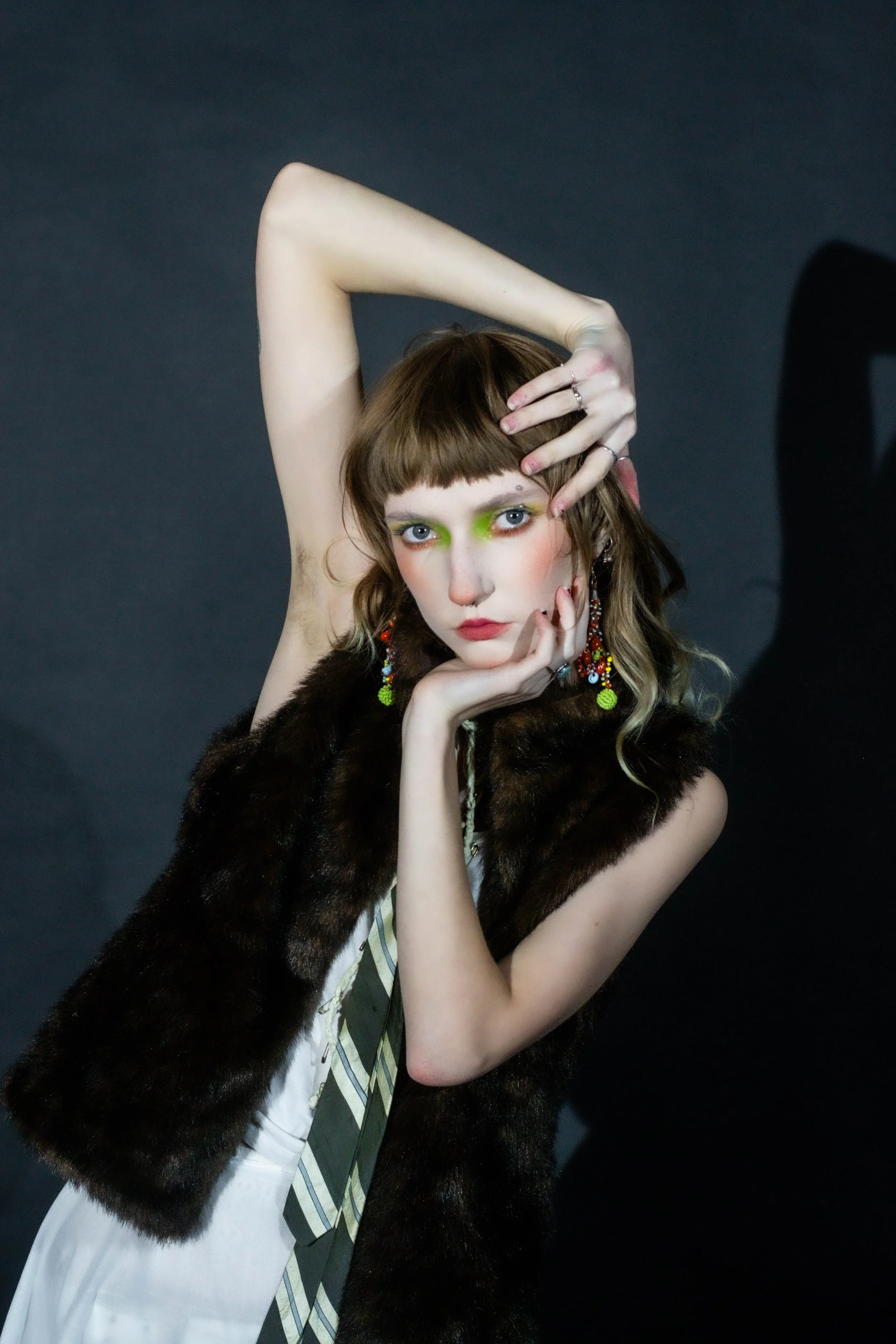 Young woman with brown hair and bangs, wearing colorful earrings, a fur vest, and a striped dress, striking a pose against a dark background.