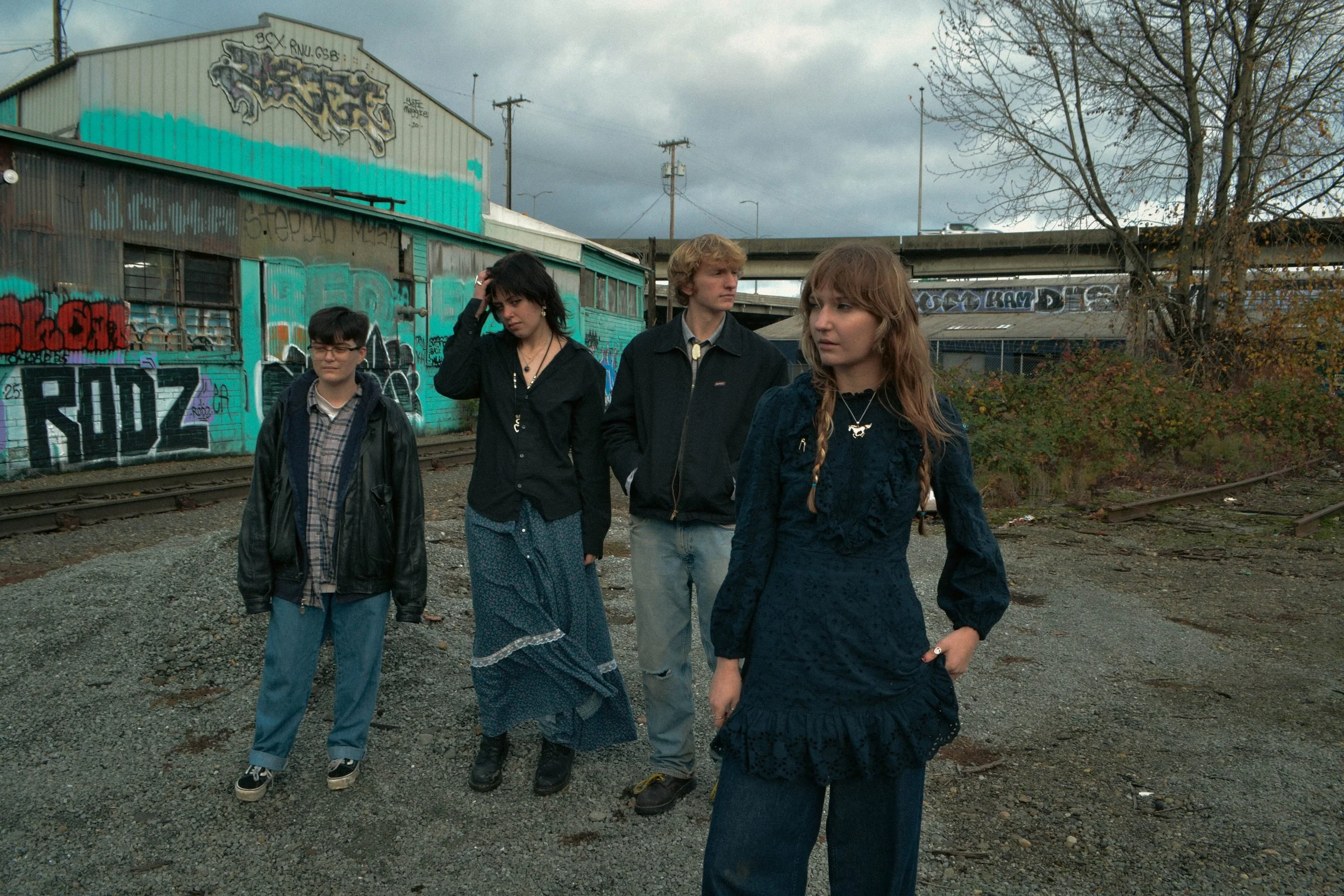 Four young people walking along railway tracks beside a graffiti-covered building on a cloudy day. The group consists of two young women and two young men, dressed casually, with trees and an overpass in the background.