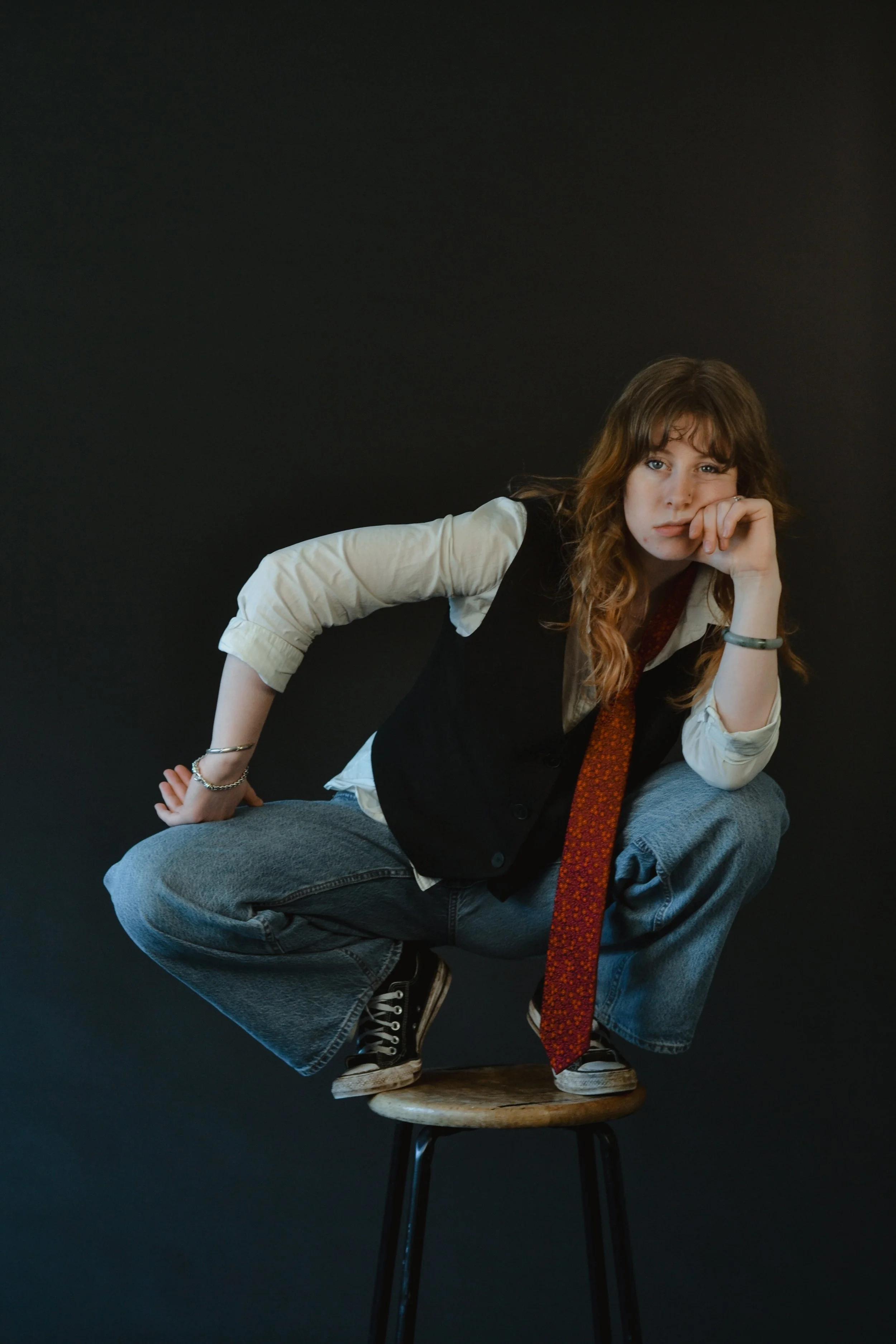 A young woman with wavy brown hair, wearing a white shirt, black vest, and loose jeans, is squatting on a wooden stool against a dark background. She rests her chin on her left hand, looking directly at the camera with a neutral expression.