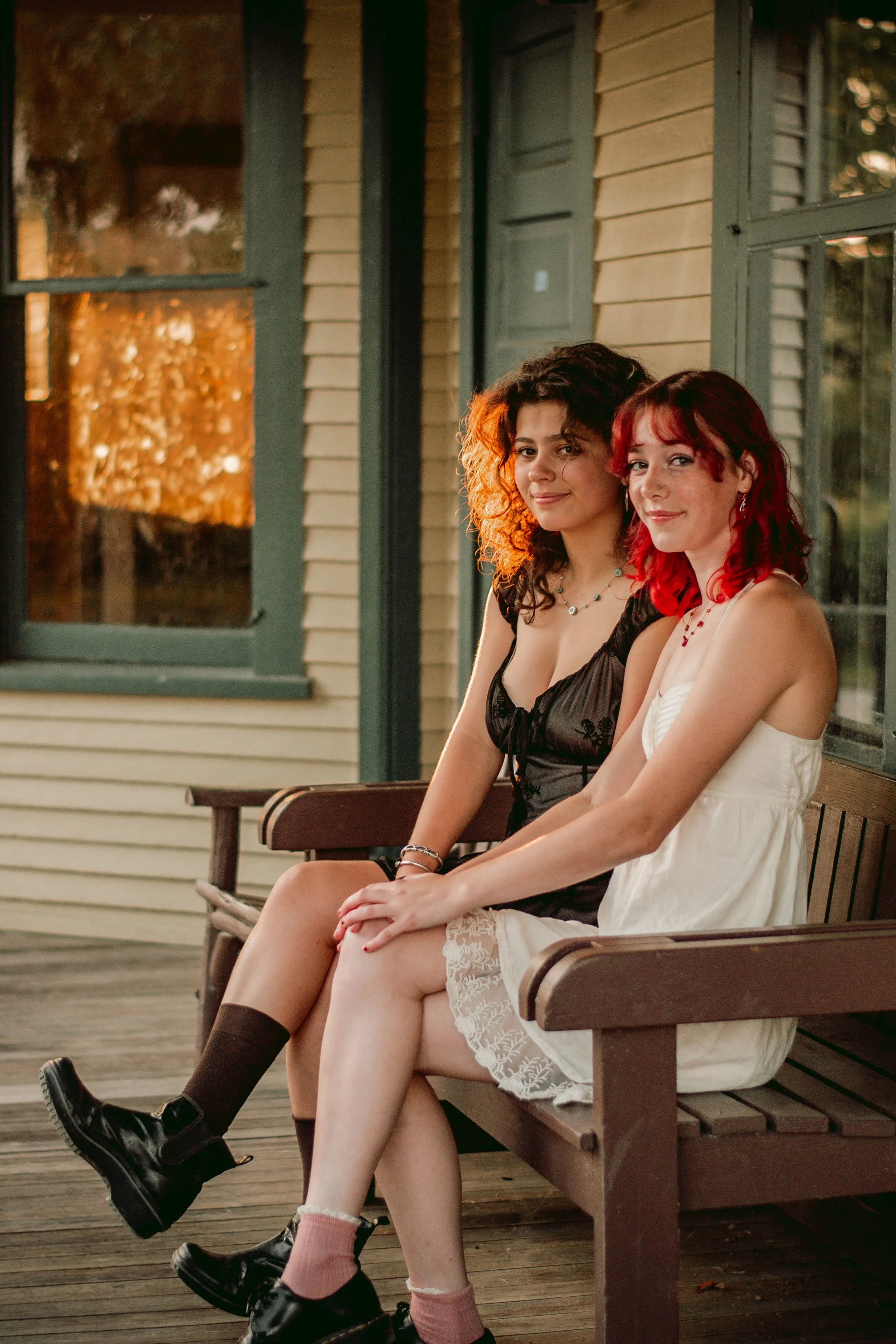 Two young women sit on a wooden bench outside a house with beige siding and green window frames, illuminated by warm sunlight, smiling.