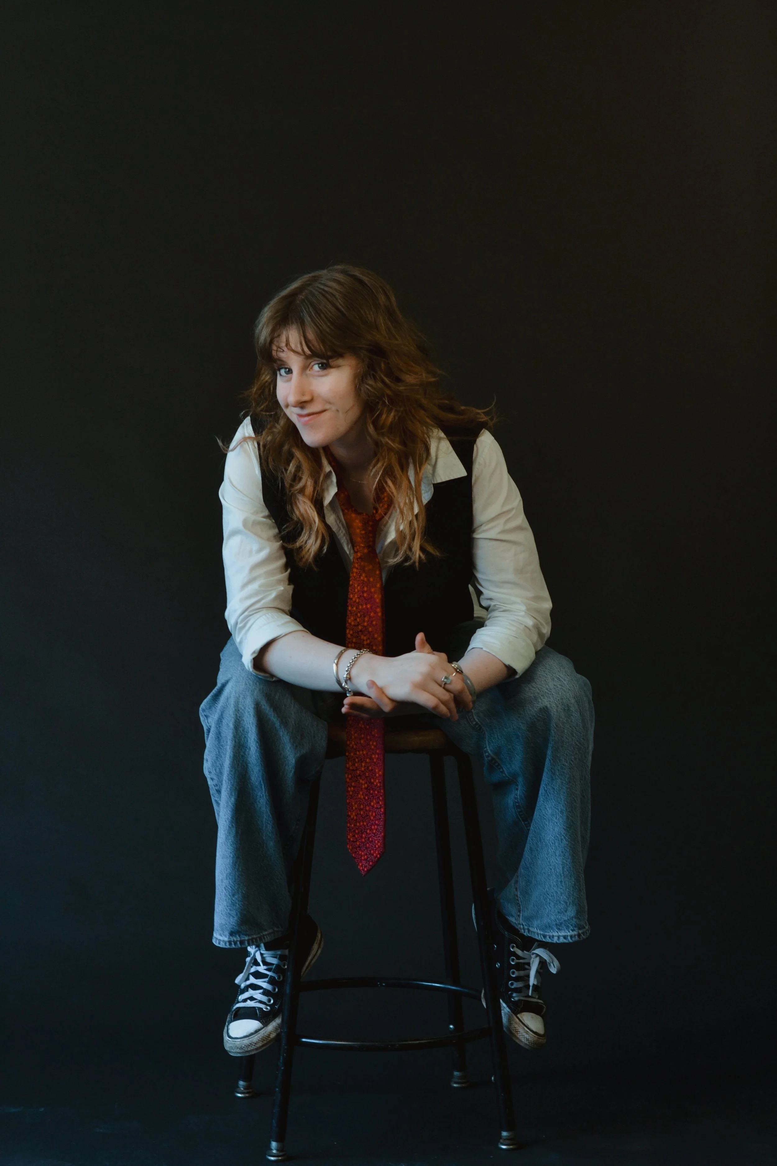 A young woman with shoulder-length wavy brown hair and light skin is sitting on a black stool against a solid black background. She is wearing a cream-colored shirt, a black vest, loose blue jeans, and black Converse sneakers. She has several bracele