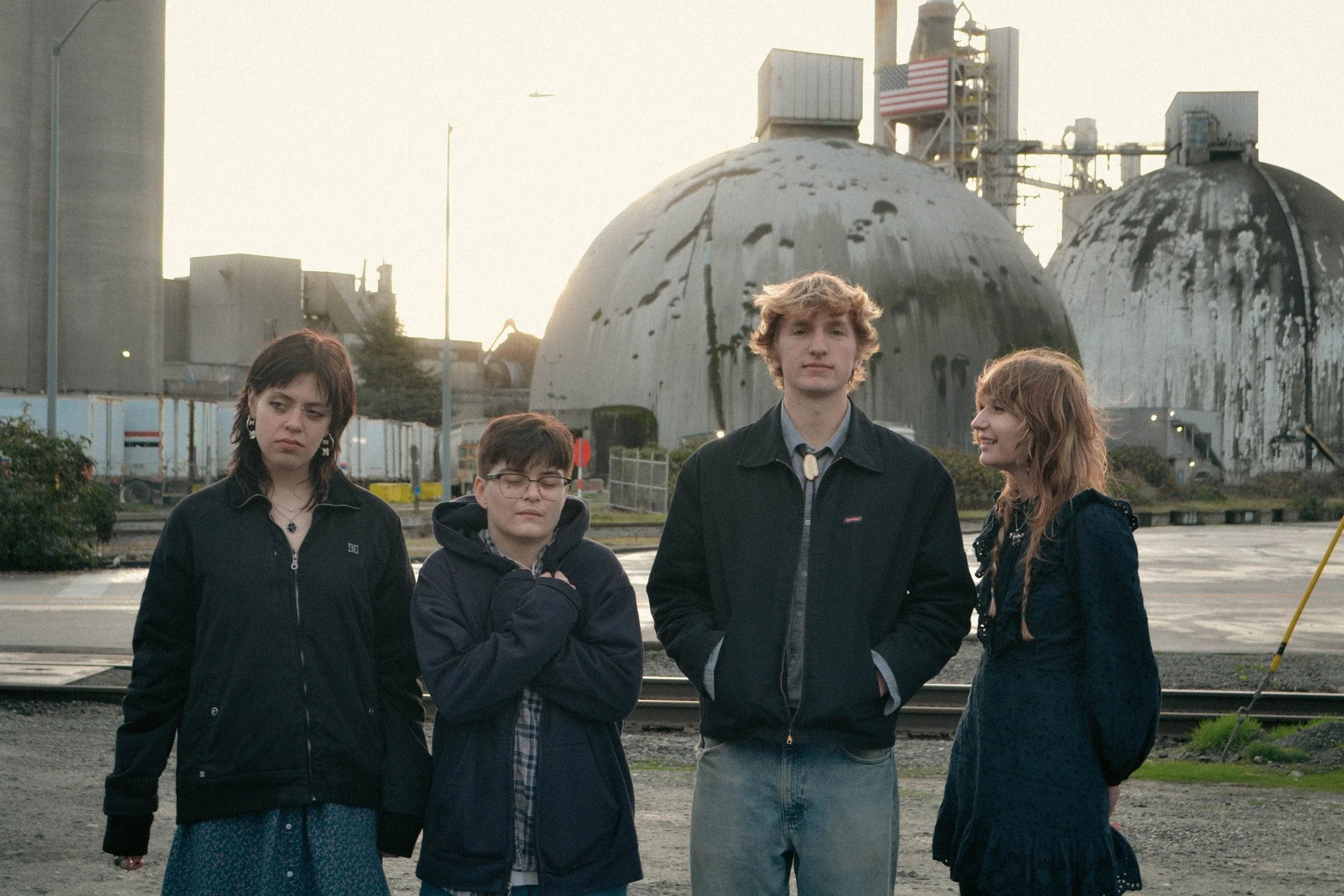Four young people standing in front of old industrial tanks outdoors, with railroad tracks visible in the background.