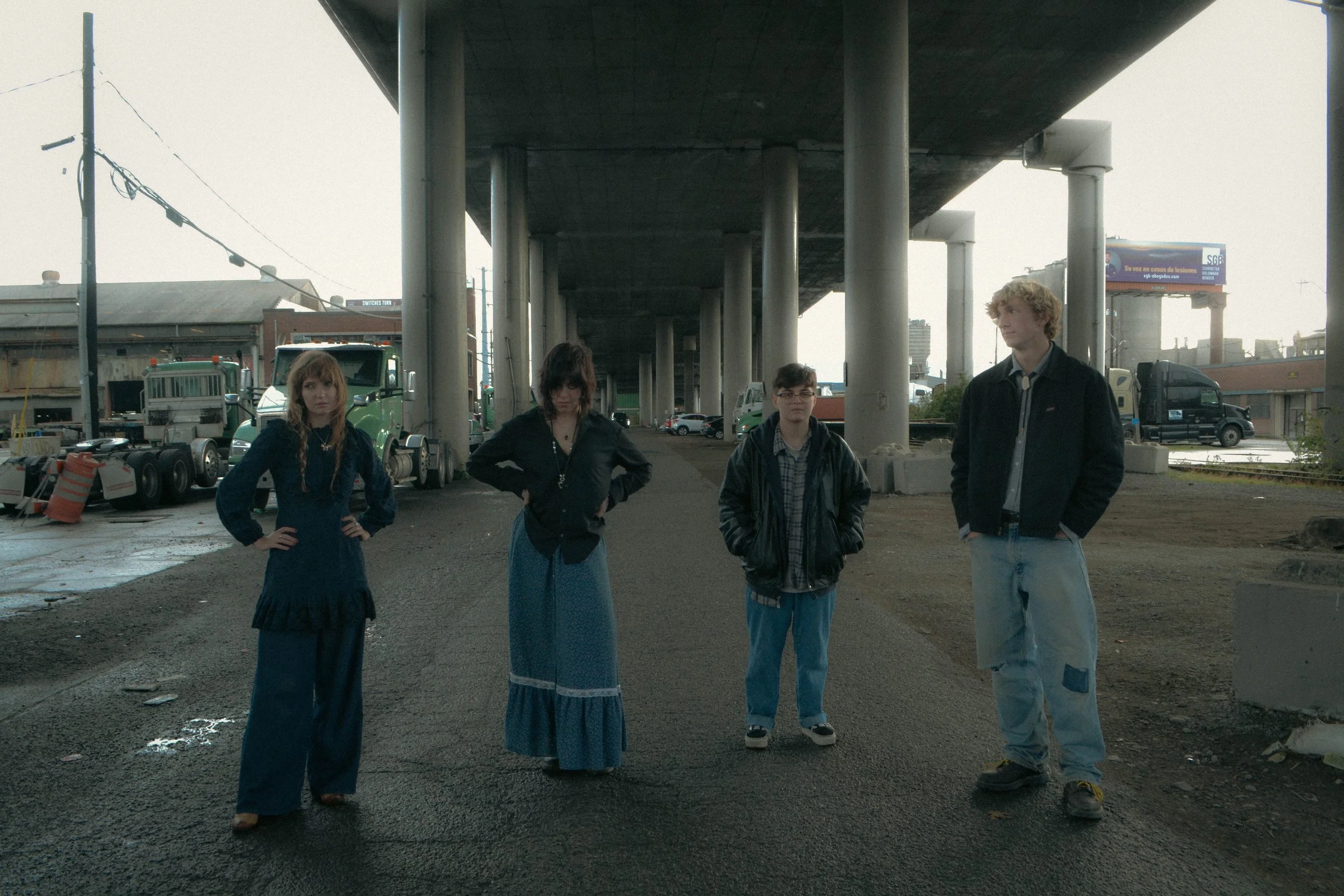 Four young individuals standing under an overpass in an urban area, with trucks and buildings in the background.