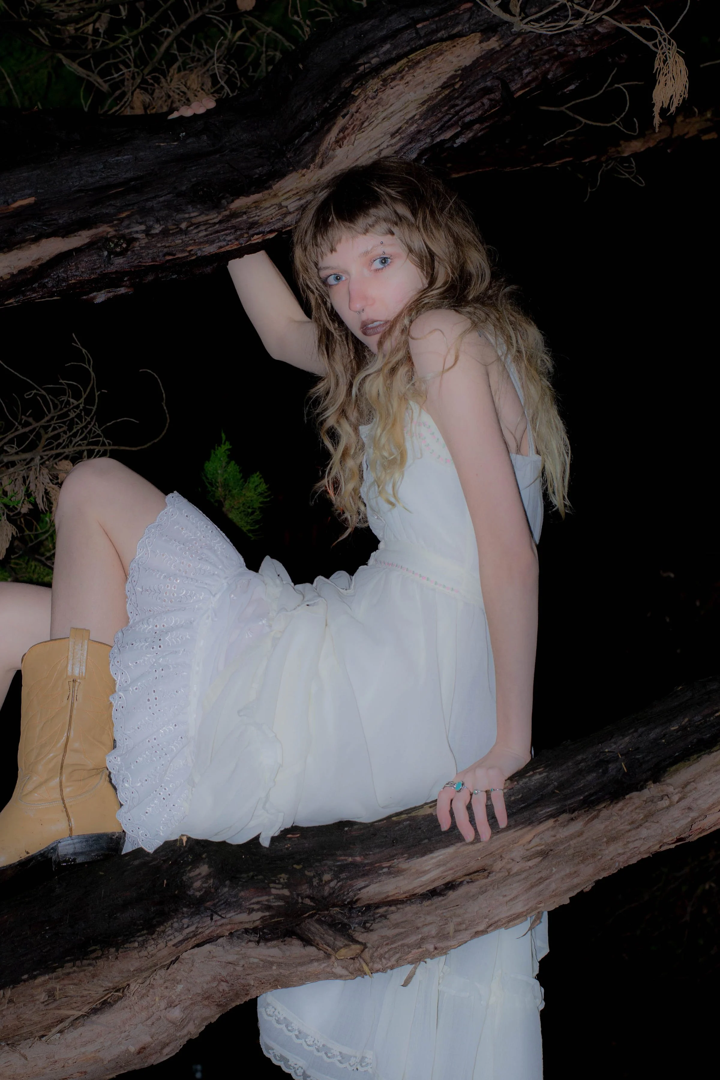 A woman with long, wavy hair, wearing a white dress and tan boots, posing amidst large fallen tree branches in a forest at night.