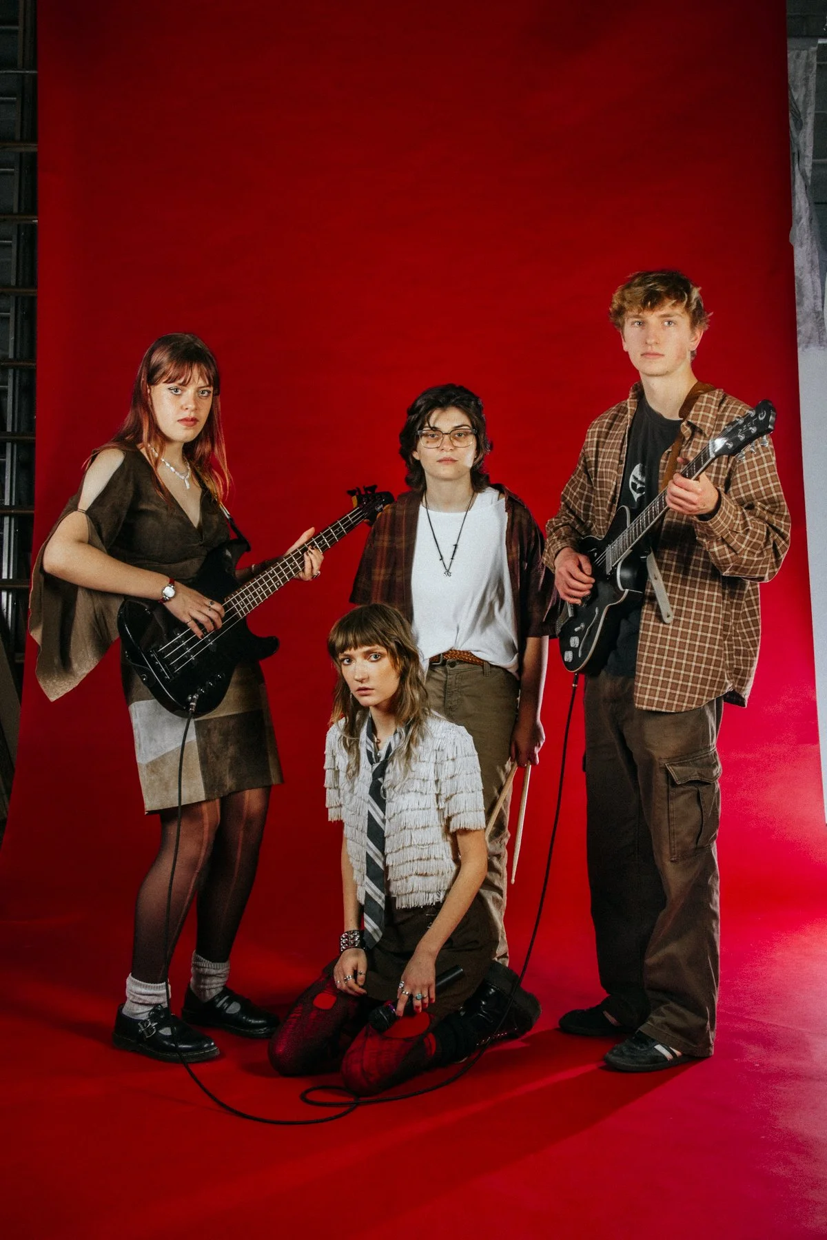 A group of five young people, four standing and one kneeling, holding musical instruments against a red backdrop. The group appears to be posing for a photo with serious expressions.